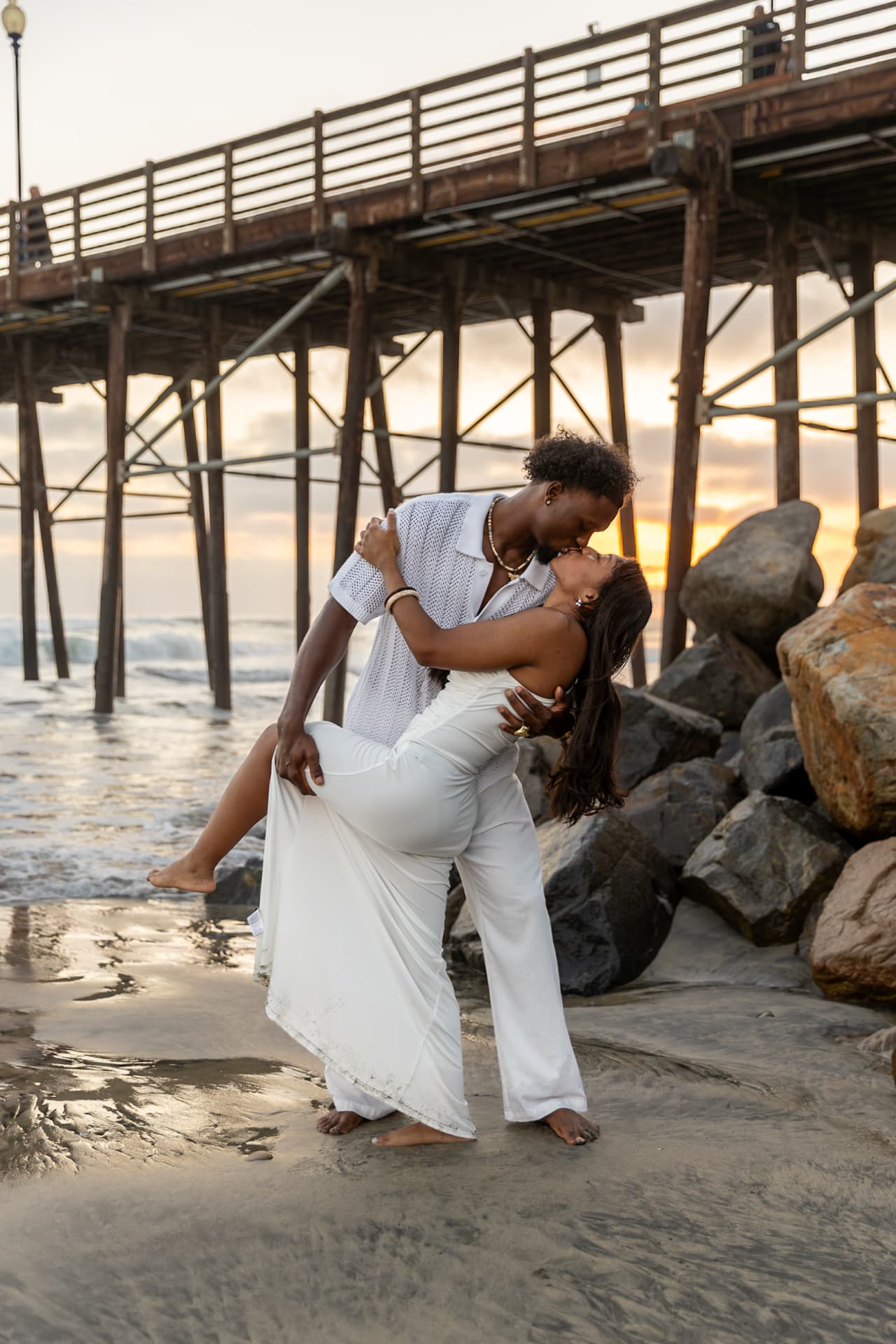Couple kissing beneath a seaside pier during golden hour