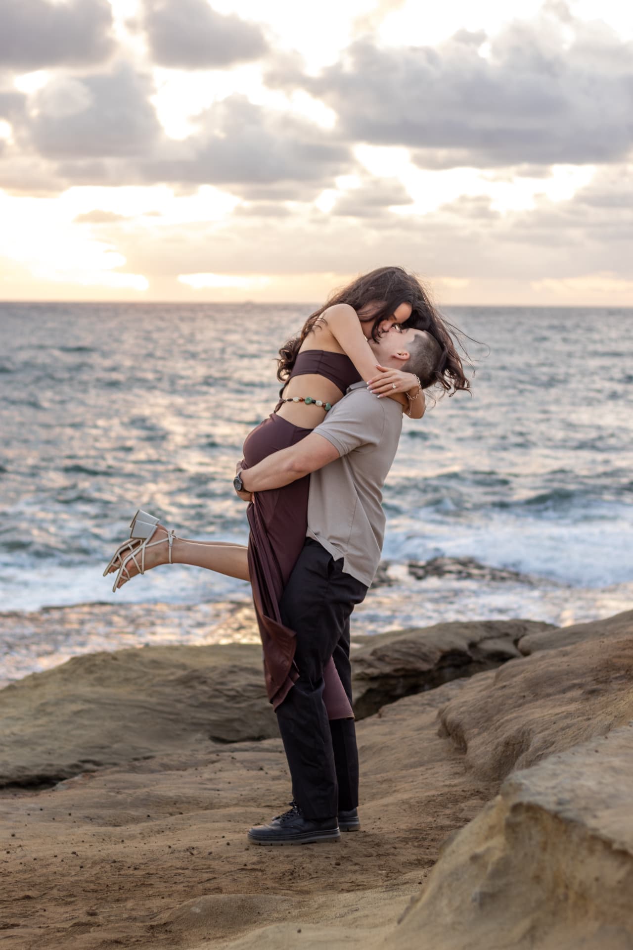 Couple holding each other on the rocks as the sun sets over the ocean