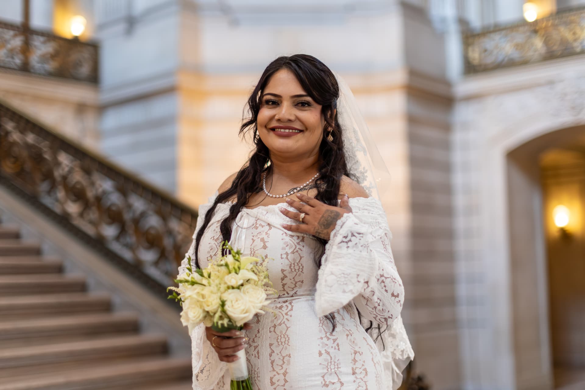 Bride smiling warmly at the camera in a grand interior, wearing a lace dress and pearl necklace, her tattooed ringed hand resting near her collarbone beside an ornate gold railing, Crystal & Joey wedding