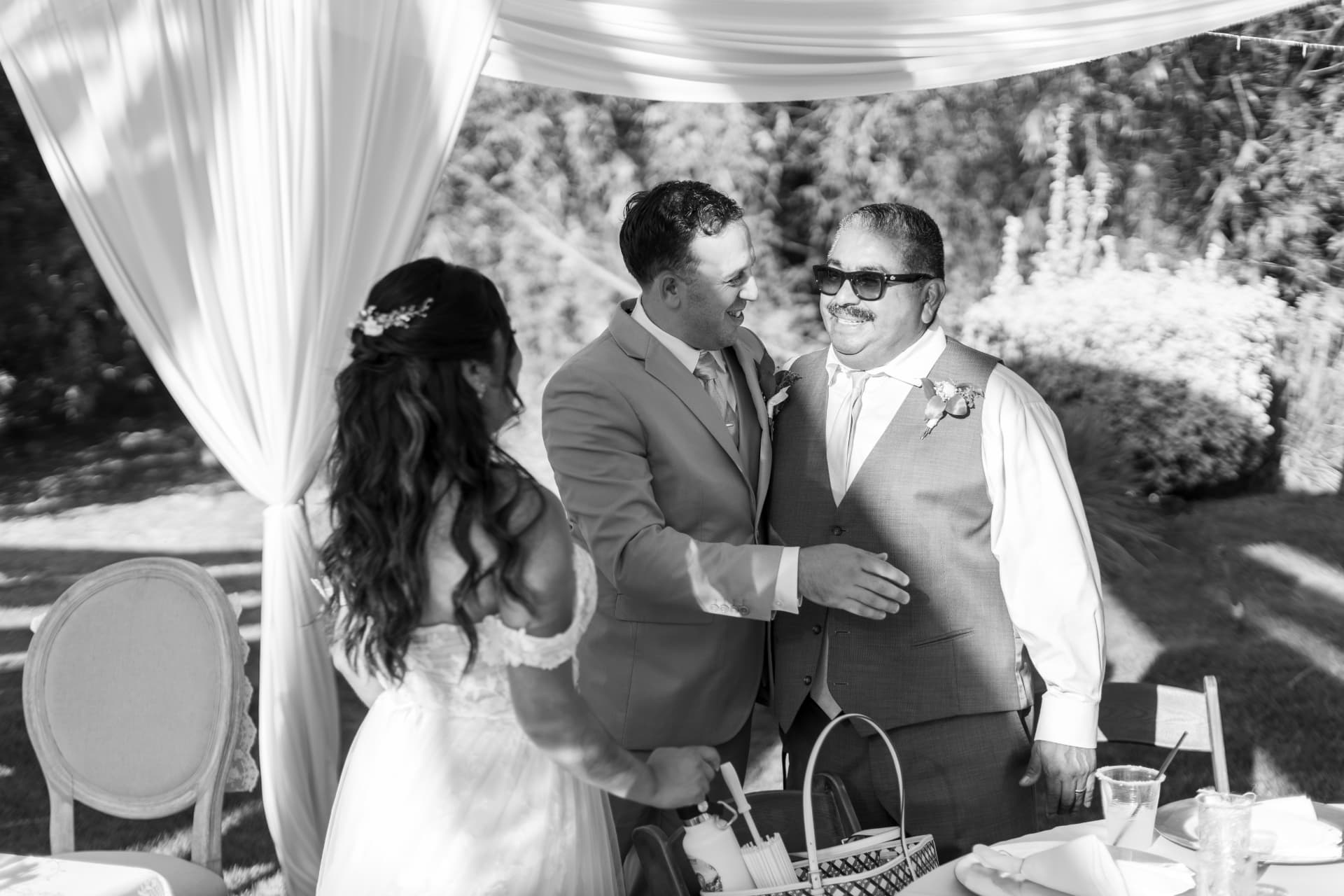 Black and white photo of a groom embracing an older man under a white draped tent as the bride watches from behind, Ashley & Sean wedding