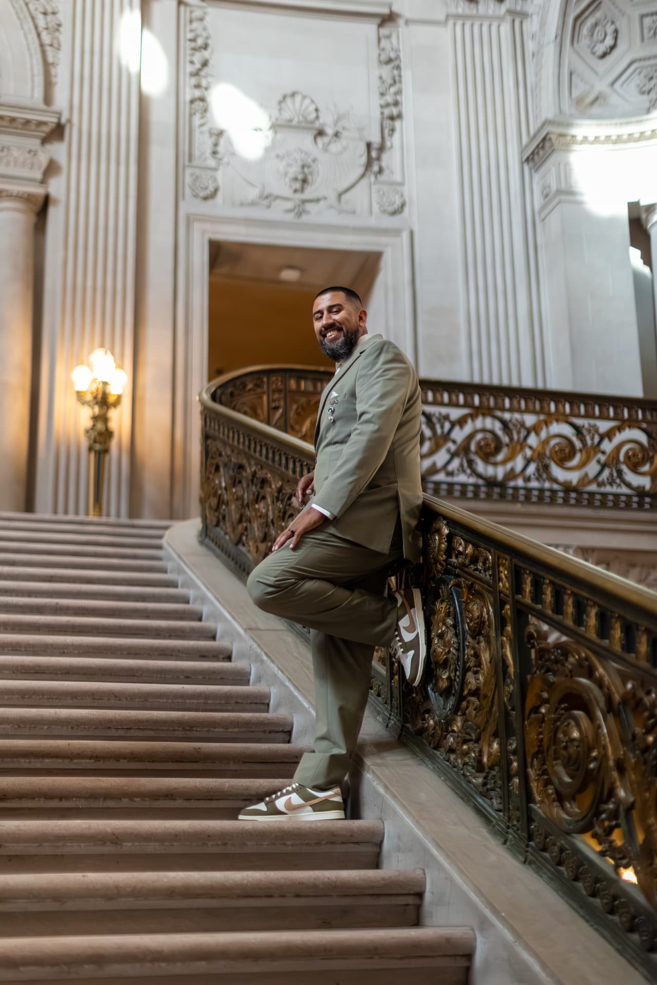 Groom in an olive-green suit and sneakers leaning back against an ornate golden railing on a grand stone staircase, smiling at the camera amid classical columns and carvings, Crystal & Joey wedding