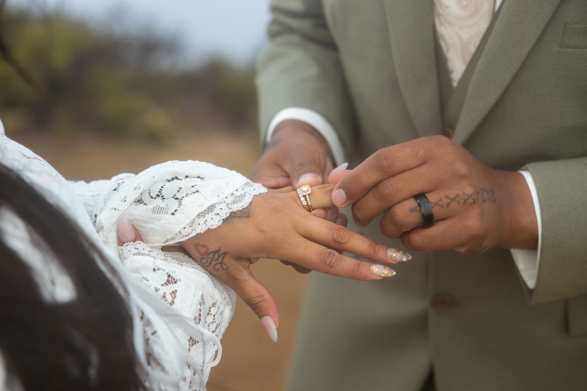 Close-up of the groom's tattooed hands placing a gold and diamond wedding ring set on the bride's finger in natural light, Crystal & Joey wedding