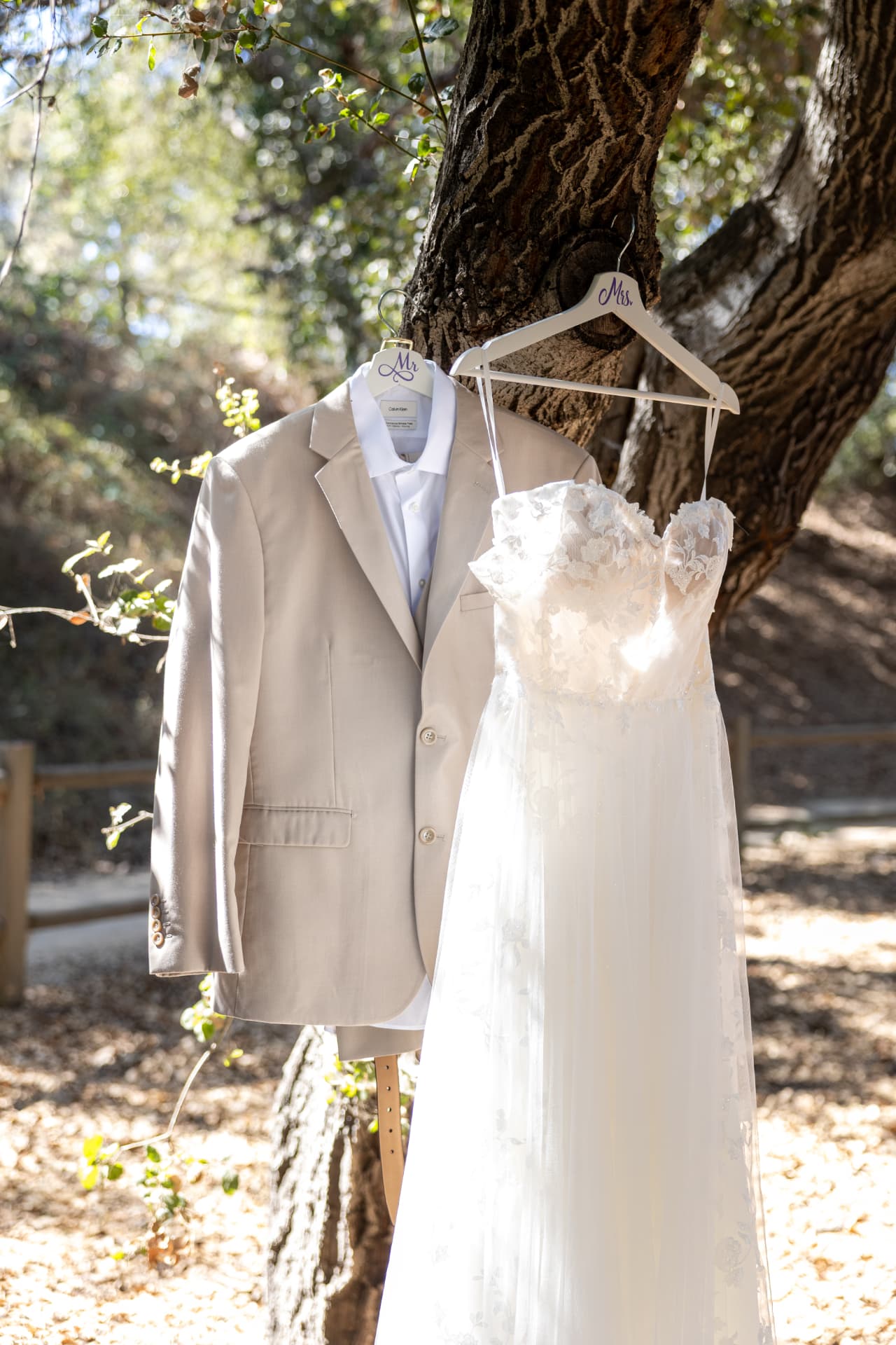 A tan suit and a white lace wedding dress hanging from a large tree in a sun-dappled wooded setting, creating a peaceful mood of anticipation for Ashley & Sean's wedding