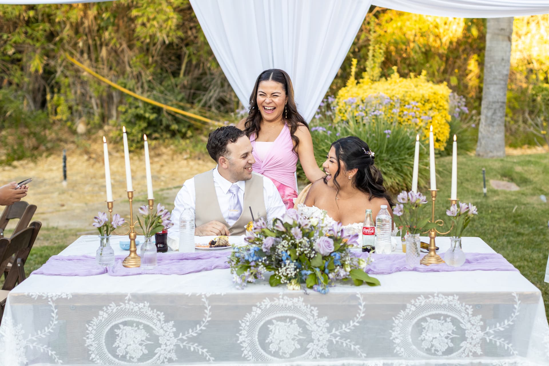 Bride and groom laughing hysterically at their sweetheart table as a guest behind them laughs along, surrounded by floral centerpieces and candles, Ashley & Sean wedding