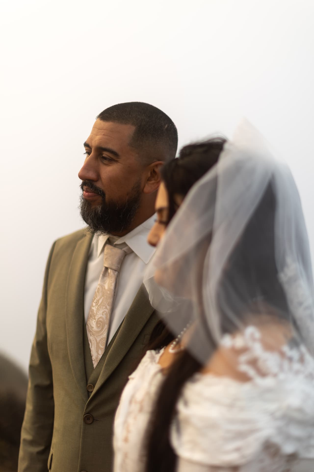 Close-up profile of the groom with a trimmed beard looking thoughtfully off-camera in a sage-green suit, the bride softly blurred beside him, Crystal & Joey wedding