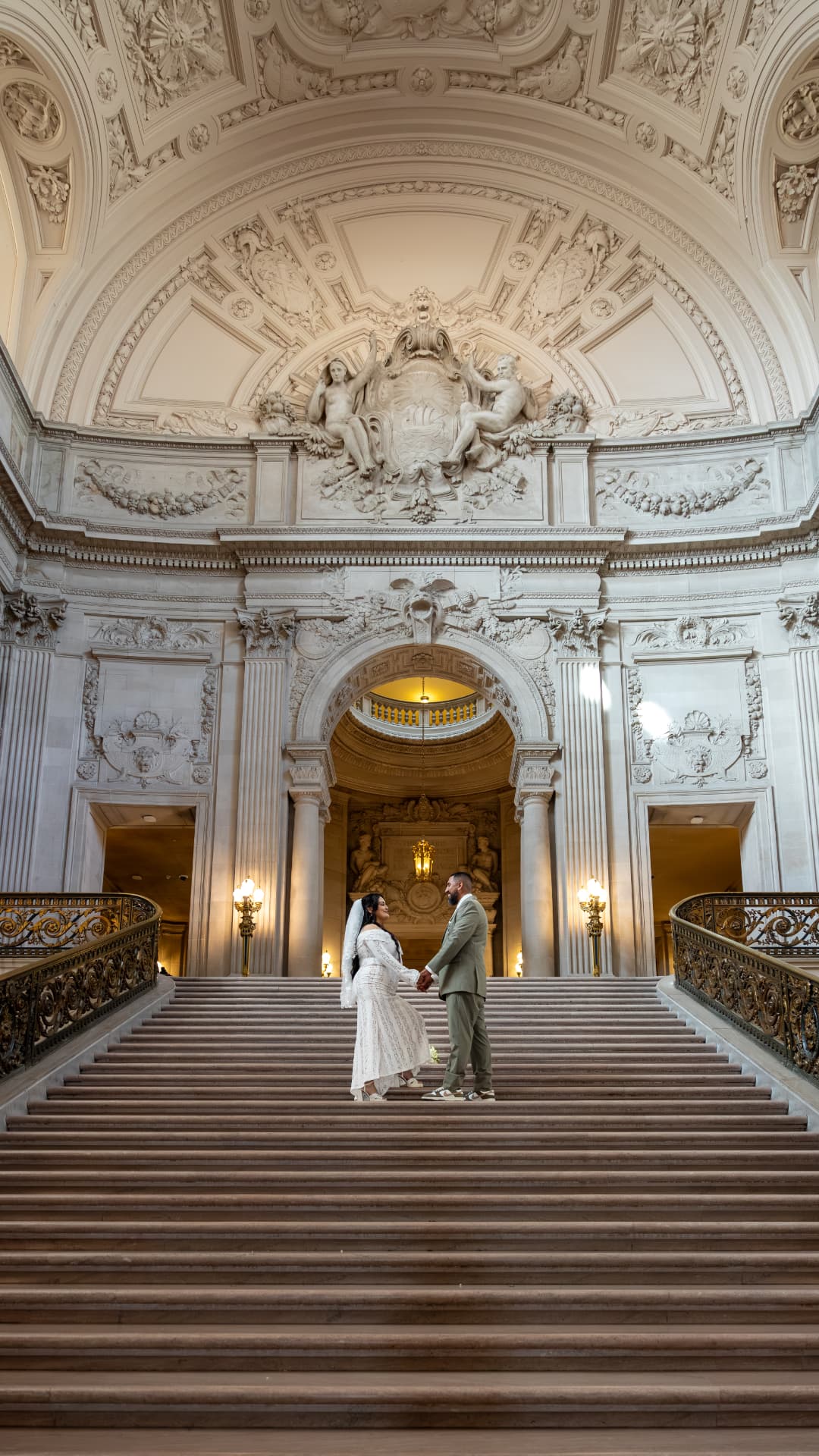 Bride in a long-sleeve lace gown and groom in a sage-green suit standing hand-in-hand on the wide marble grand staircase of a majestic classical building, Crystal & Joey wedding