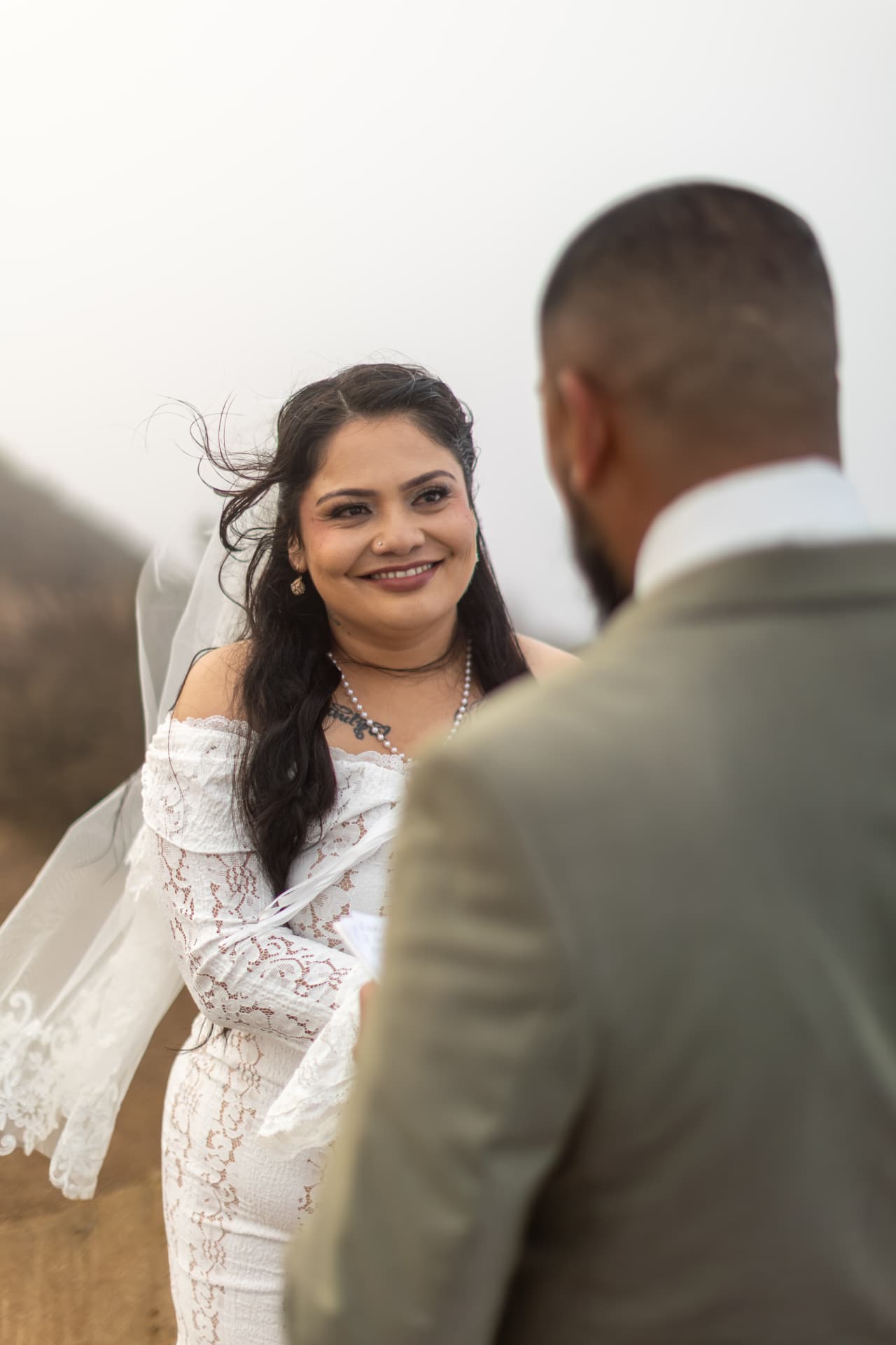 Bride with dark hair blowing in the breeze smiling warmly at her groom, wearing a pearl necklace and off-the-shoulder lace dress against misty brown hills, Crystal & Joey wedding