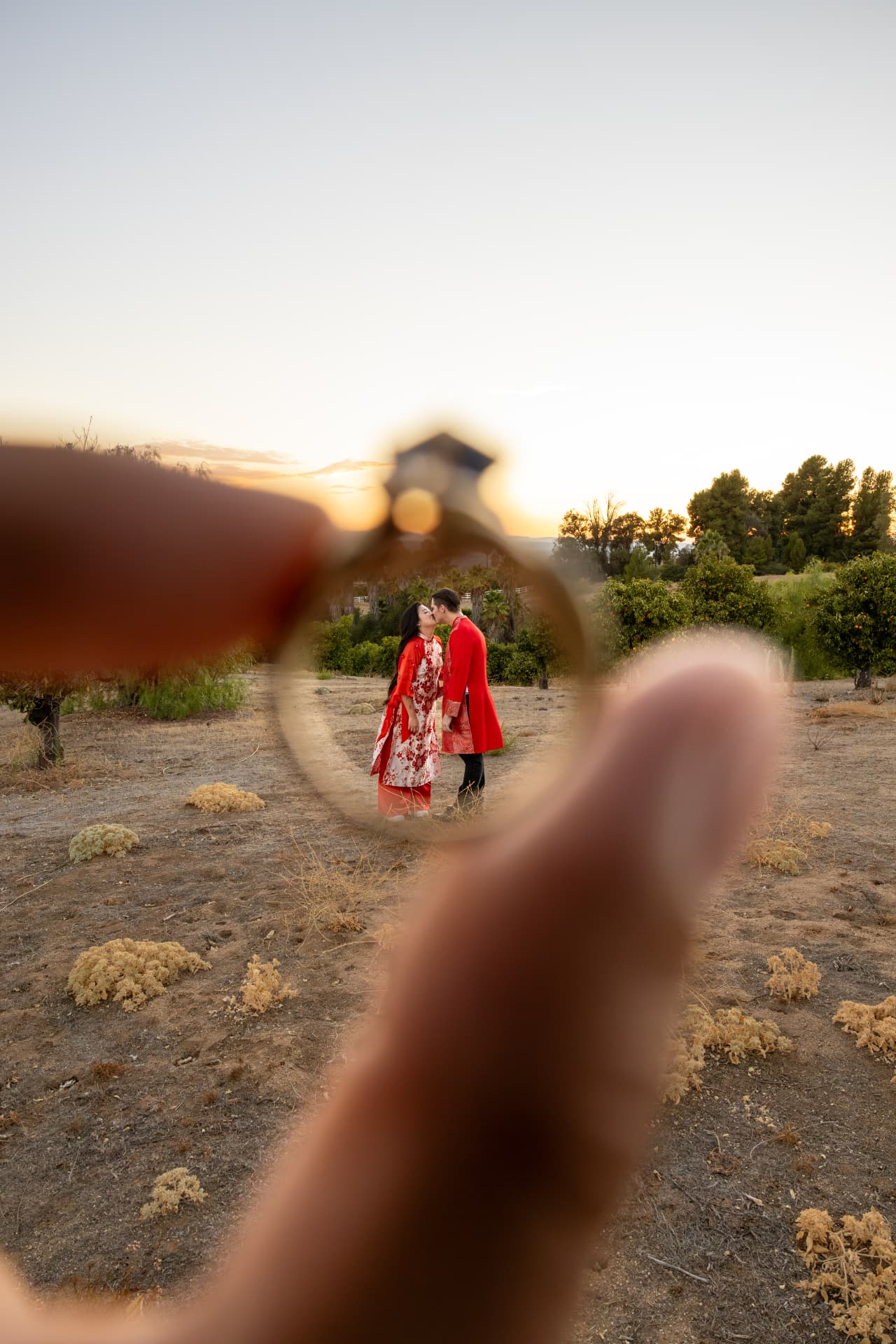 Couple in red traditional Vietnamese clothing kissing in a dry open field at sunset, framed through an engagement ring held in the foreground, Thanh & Nicholas engagement