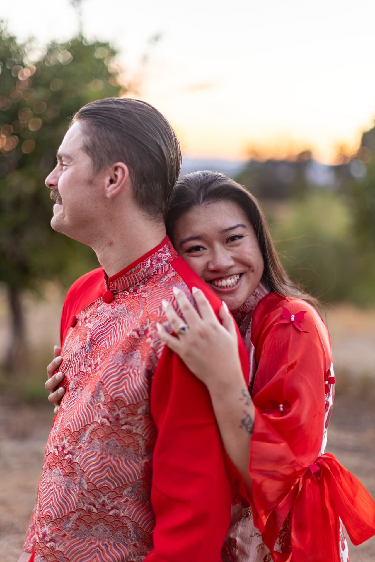 Woman smiling warmly at the camera while embracing a man from behind in golden sunset light, Thanh & Nicholas engagement
