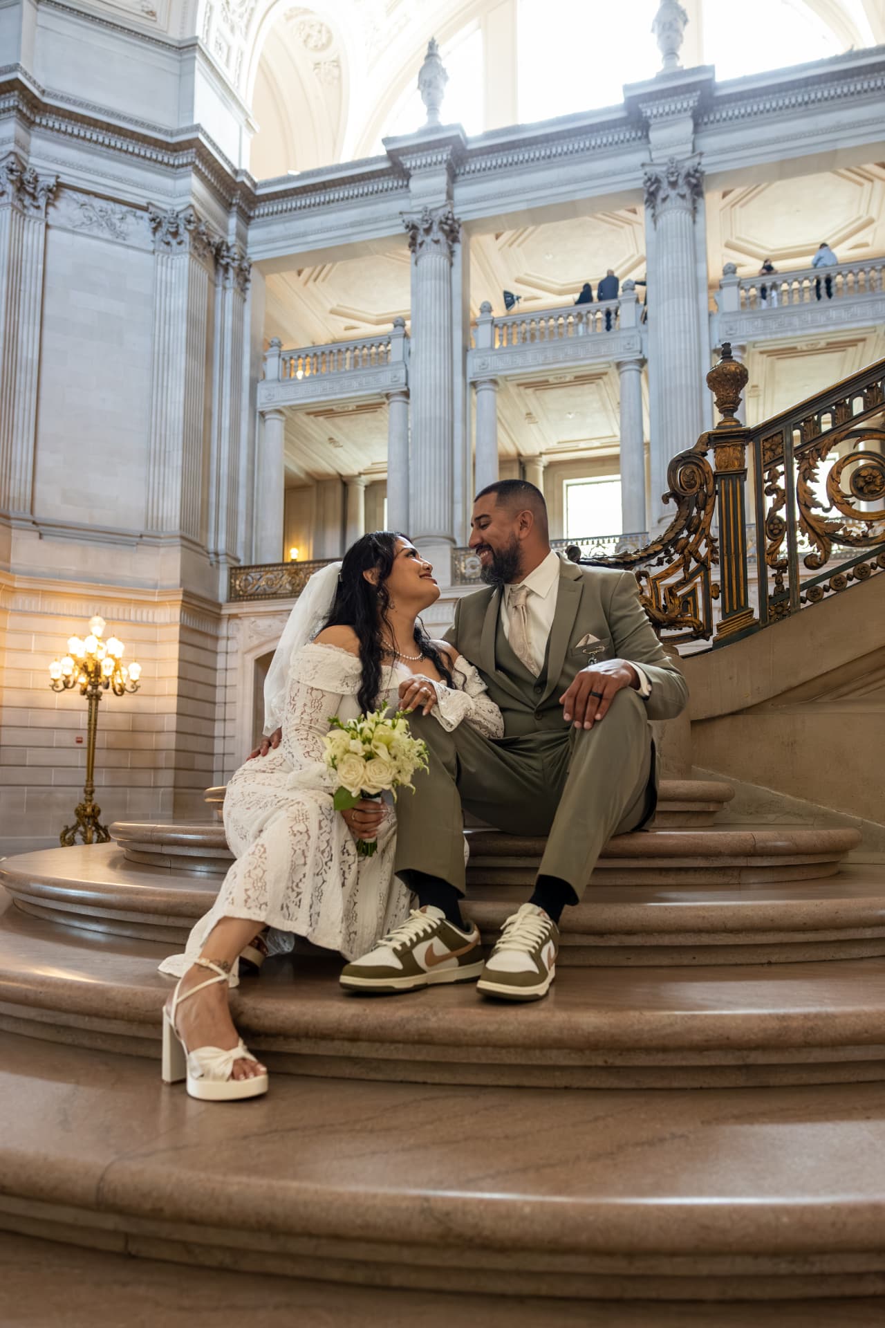 Bride and groom sitting together on the lower steps of a grand ornate staircase, smiling at each other, the bride in white heels and the groom in sage-green with sneakers, Crystal & Joey wedding