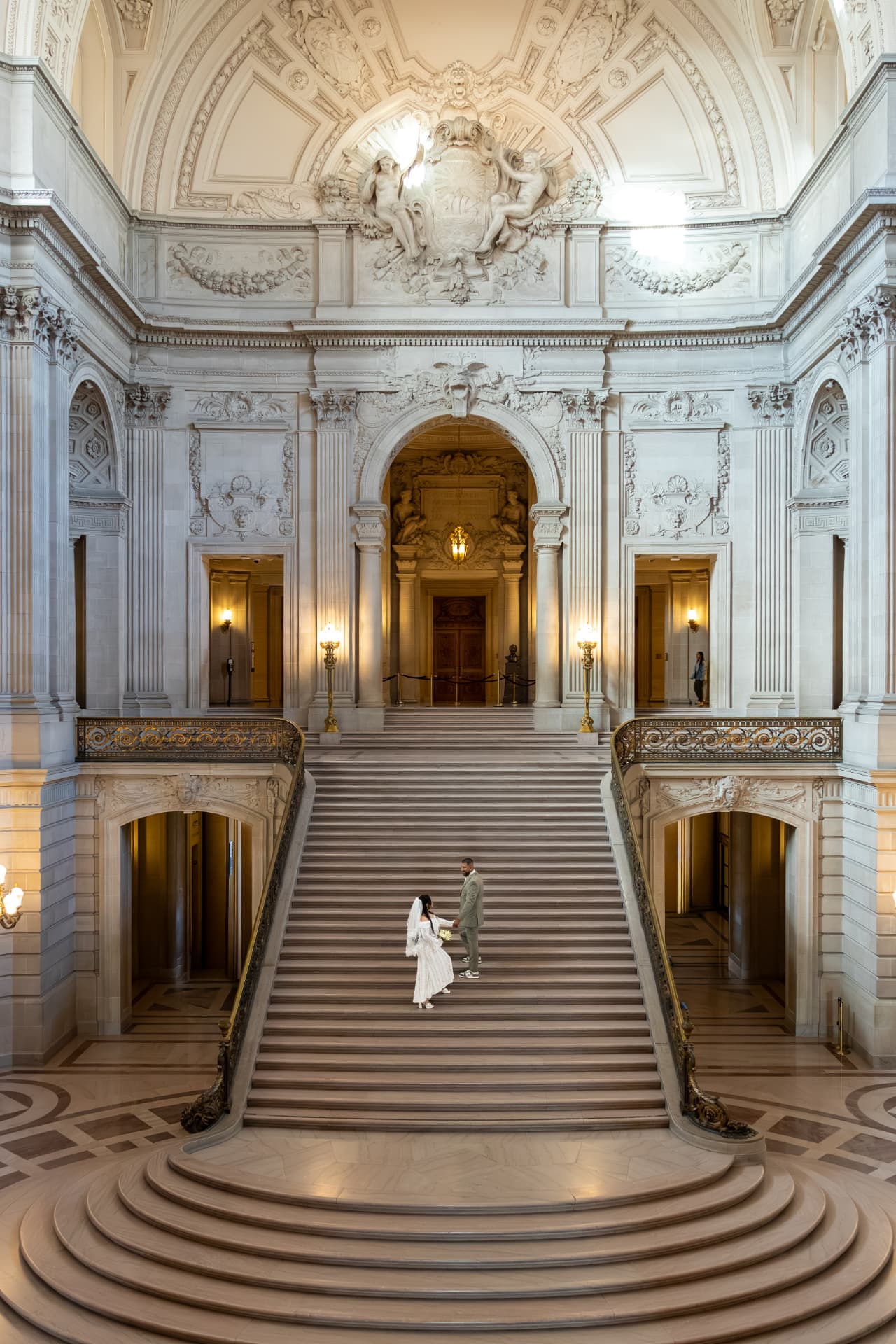 Bride and groom holding hands in the center of a massive symmetrical marble staircase within a grand warmly lit classical hall, Crystal & Joey wedding