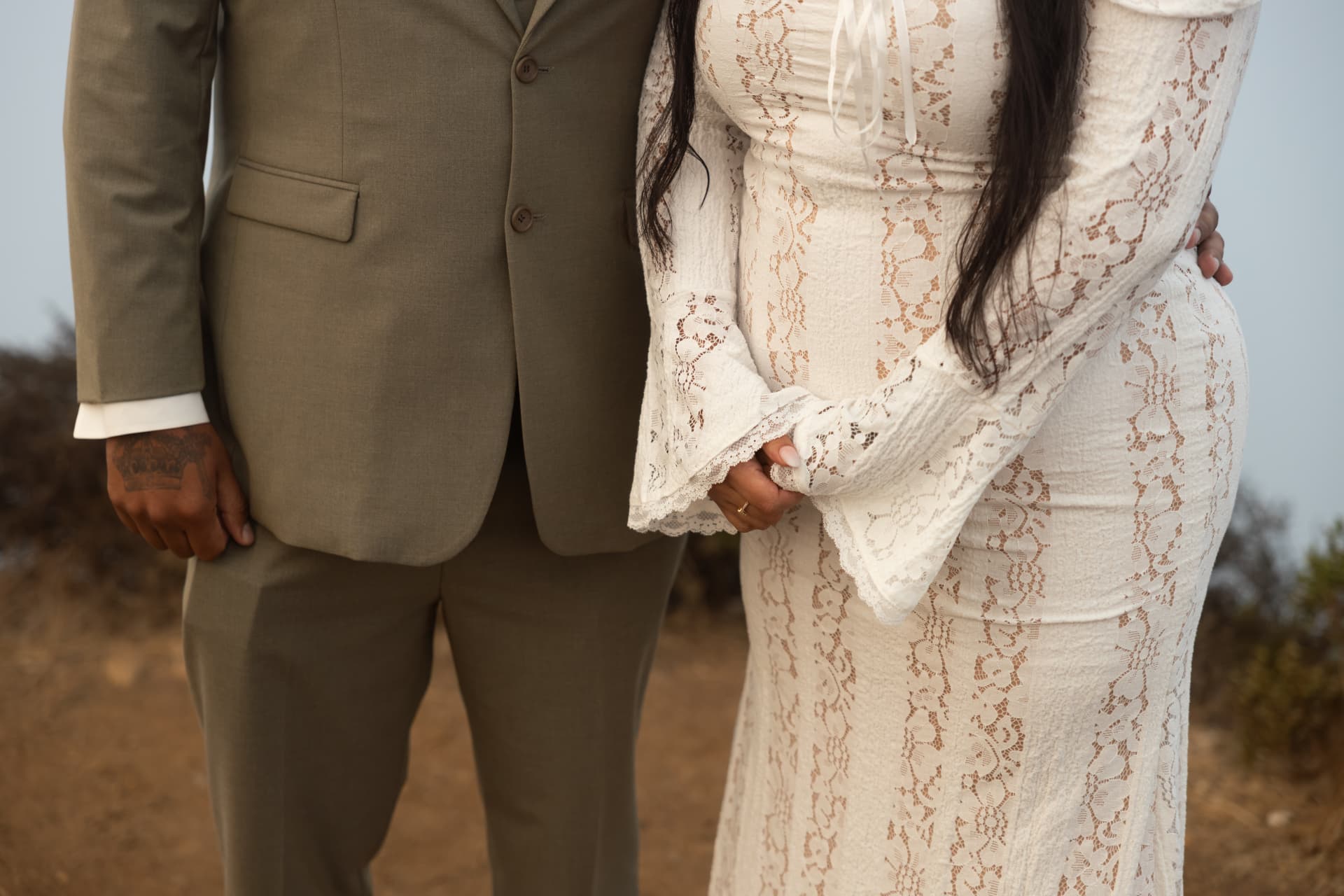 Groom in an olive-green suit and bride in a white lace dress smiling at each other against a bright hazy sky, Crystal & Joey wedding