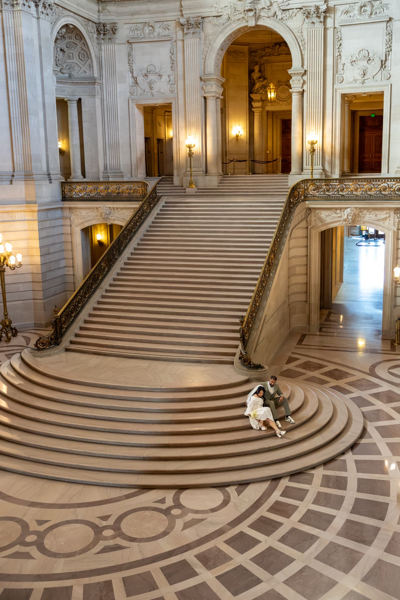 Bride and groom sitting together on the wide lower steps of a grand marble staircase viewed from above in a warmly lit patterned hall, Crystal & Joey wedding