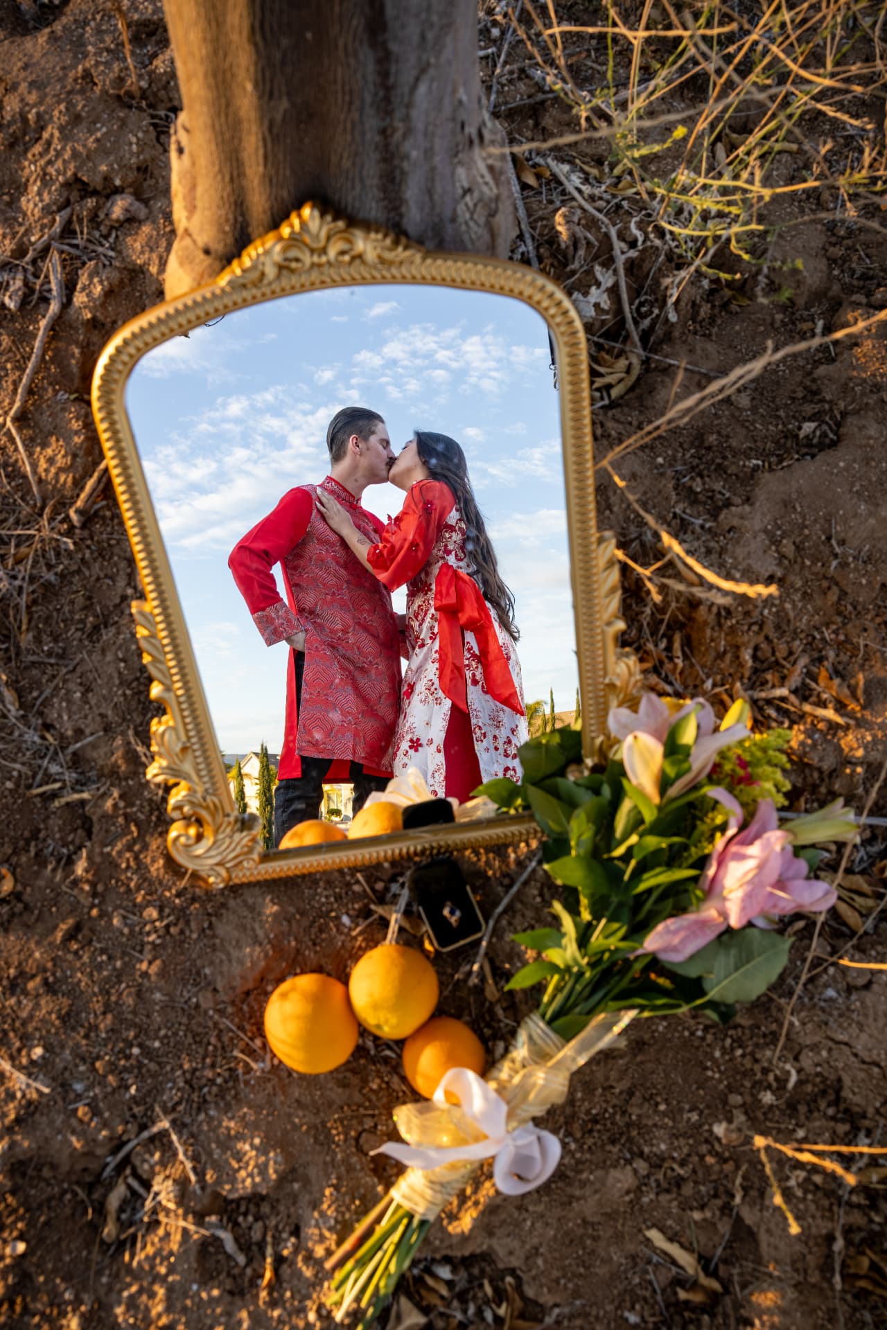 Couple in red traditional Vietnamese attire kissing in the reflection of an ornate gold mirror on dry earth with scattered oranges and lilies, Thanh & Nicholas engagement