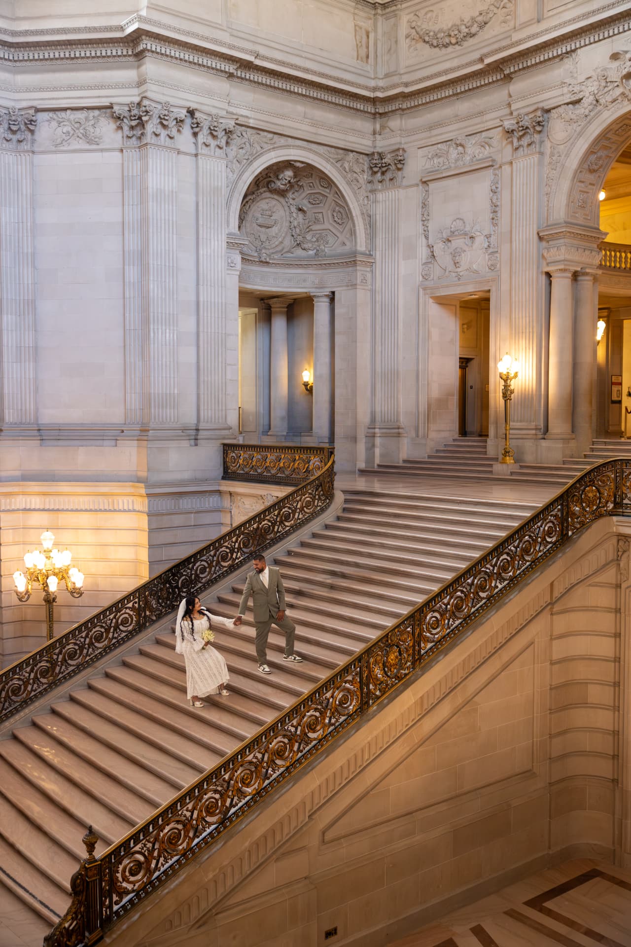 Bride and groom holding hands while ascending a grand ornate marble staircase illuminated by warm indoor lamps, Crystal & Joey wedding