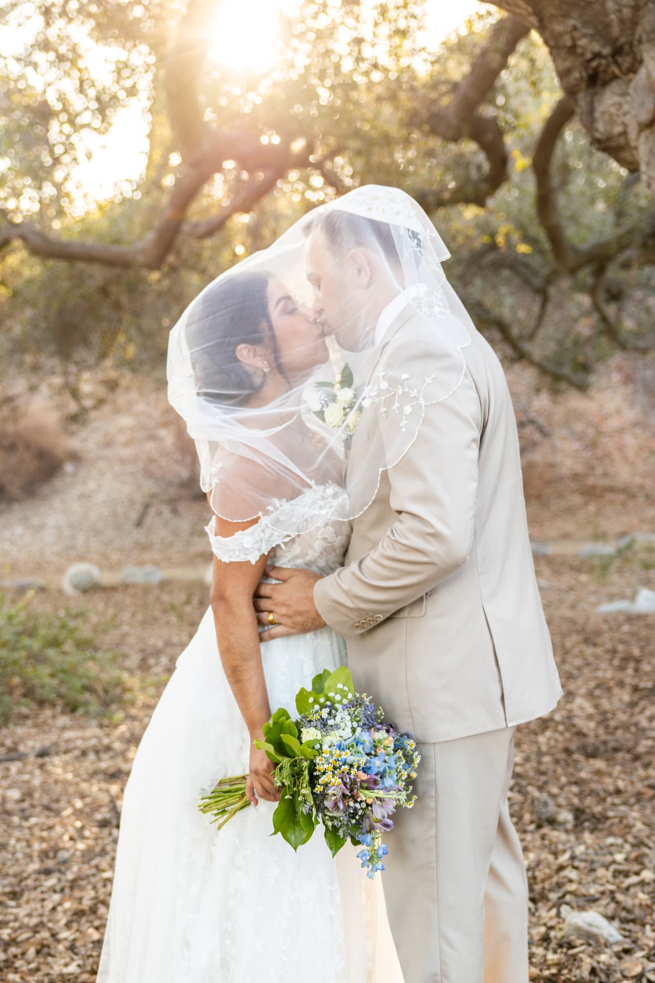 Bride and groom kissing under the veil as sunlight bursts through the oak trees