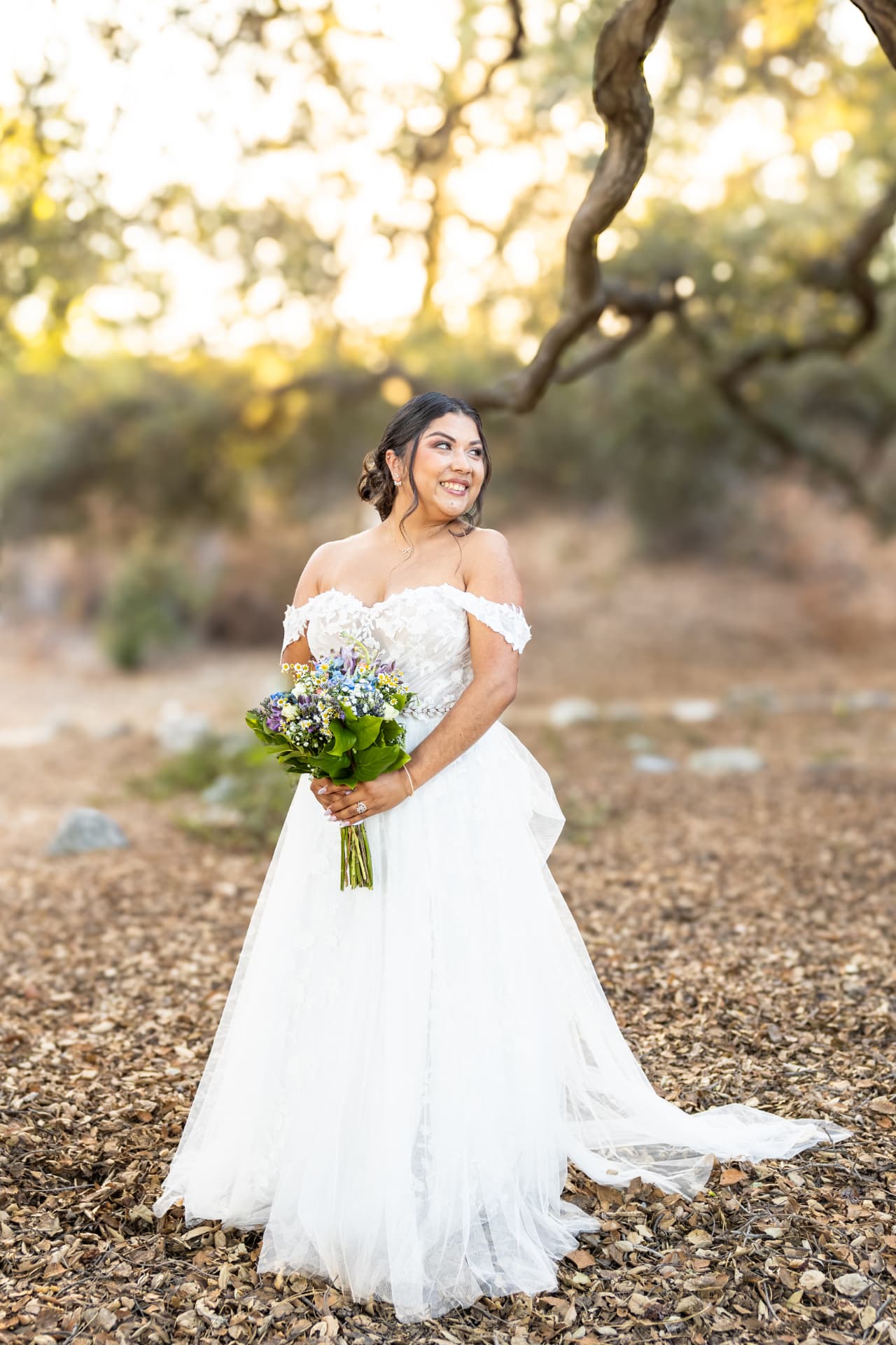 Bride in an off-shoulder lace gown holding a blue and purple wildflower bouquet in golden light