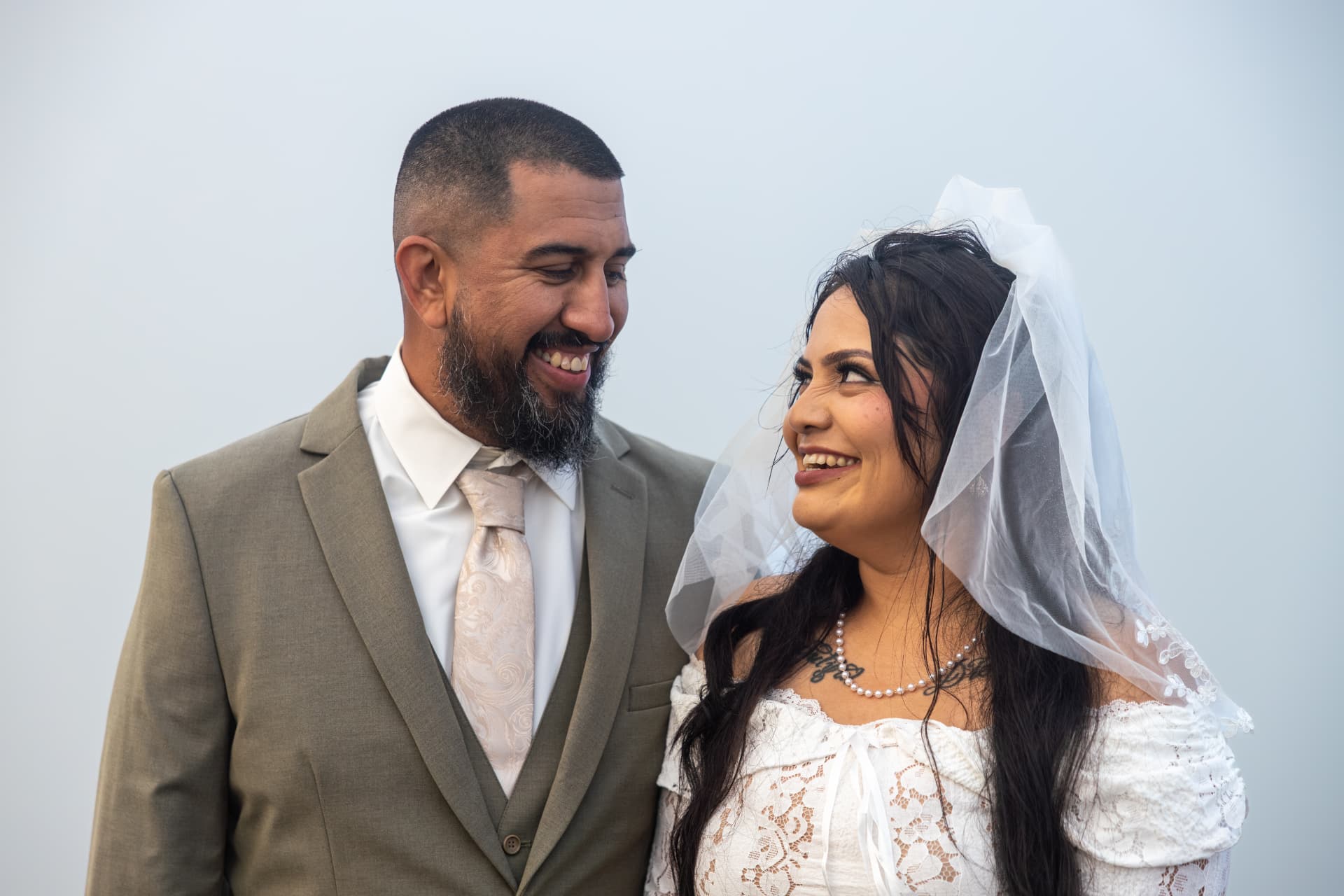Groom in an olive-green suit with his arm around a bride in a long-sleeved white lace dress on a dirt path, Crystal & Joey wedding