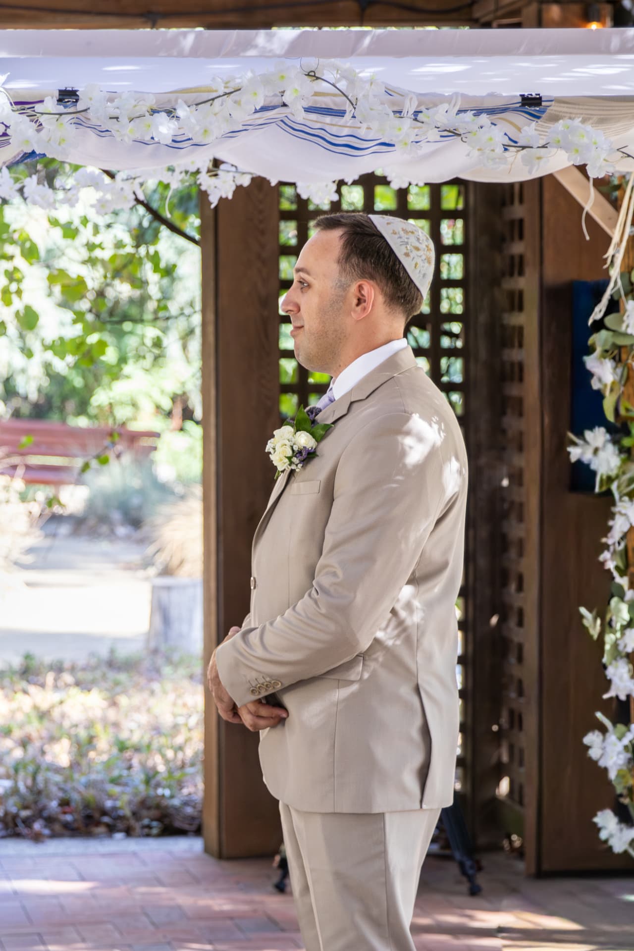 Sean standing with a hopeful expression in his tan wedding suit under a decorated chuppah in a sun-dappled outdoor garden, Ashley & Sean wedding