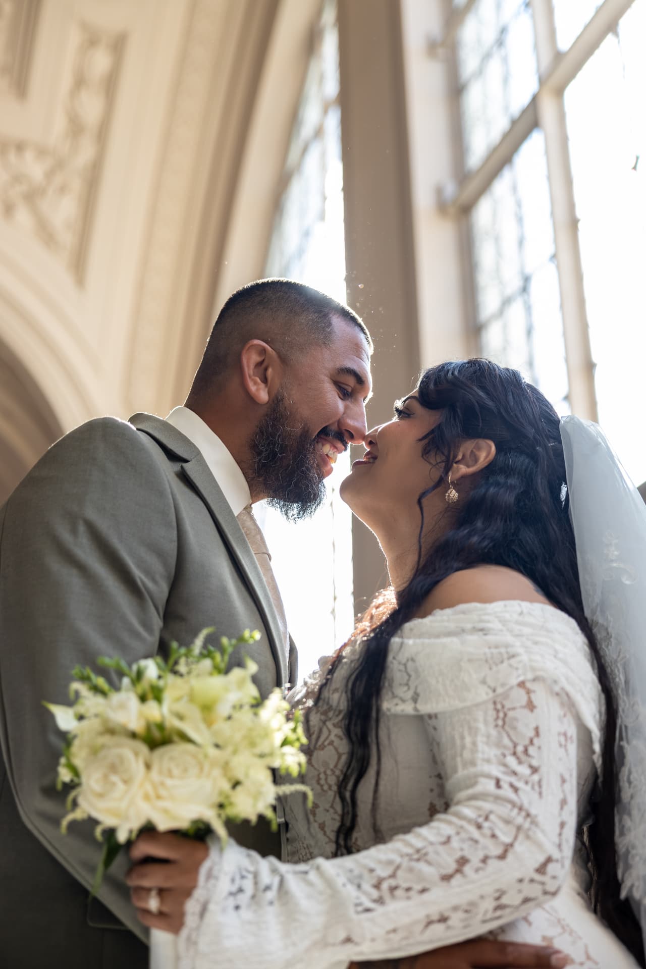 Close-up profile of bride and groom smiling with noses touching as soft window light glows behind them, the bride holding a white rose bouquet, Crystal & Joey wedding