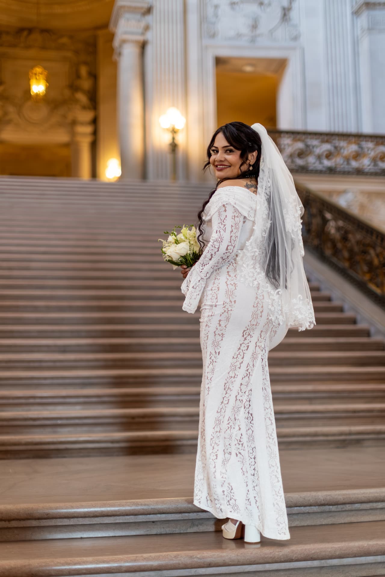Bride in a white lace gown and veil looking back over her shoulder with a smile at the base of a grand stone staircase with tall columns and warm glowing lamps, Crystal & Joey wedding