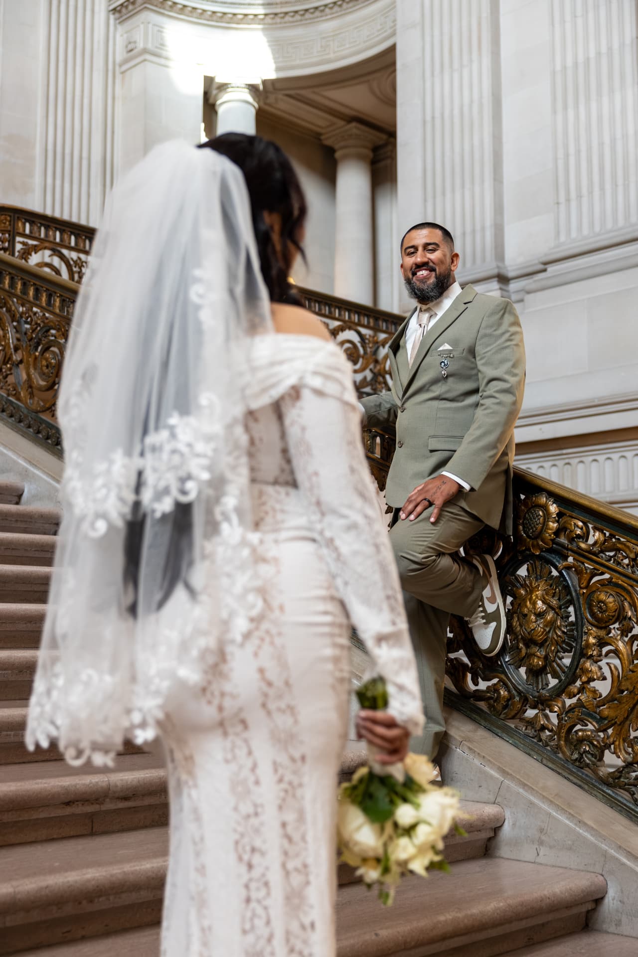 Groom in a sage-green suit smiling warmly at his veiled bride while leaning against an ornate brass railing on the marble stairs, Crystal & Joey wedding