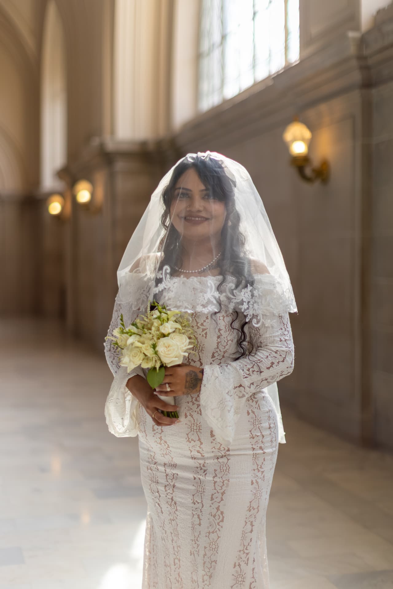 Bride centered in a sunlit grand hallway with arched ceilings, wearing a white lace dress and sheer veil and holding a small bouquet, looking softly toward the camera, Crystal & Joey wedding