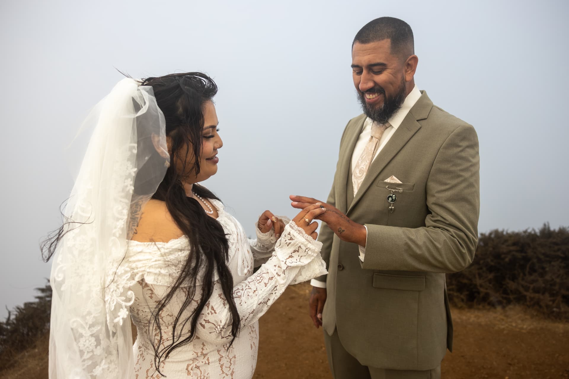 Bride smiling as she slides a wedding ring onto the groom's finger during their outdoor ceremony under a soft overcast sky, Crystal & Joey wedding