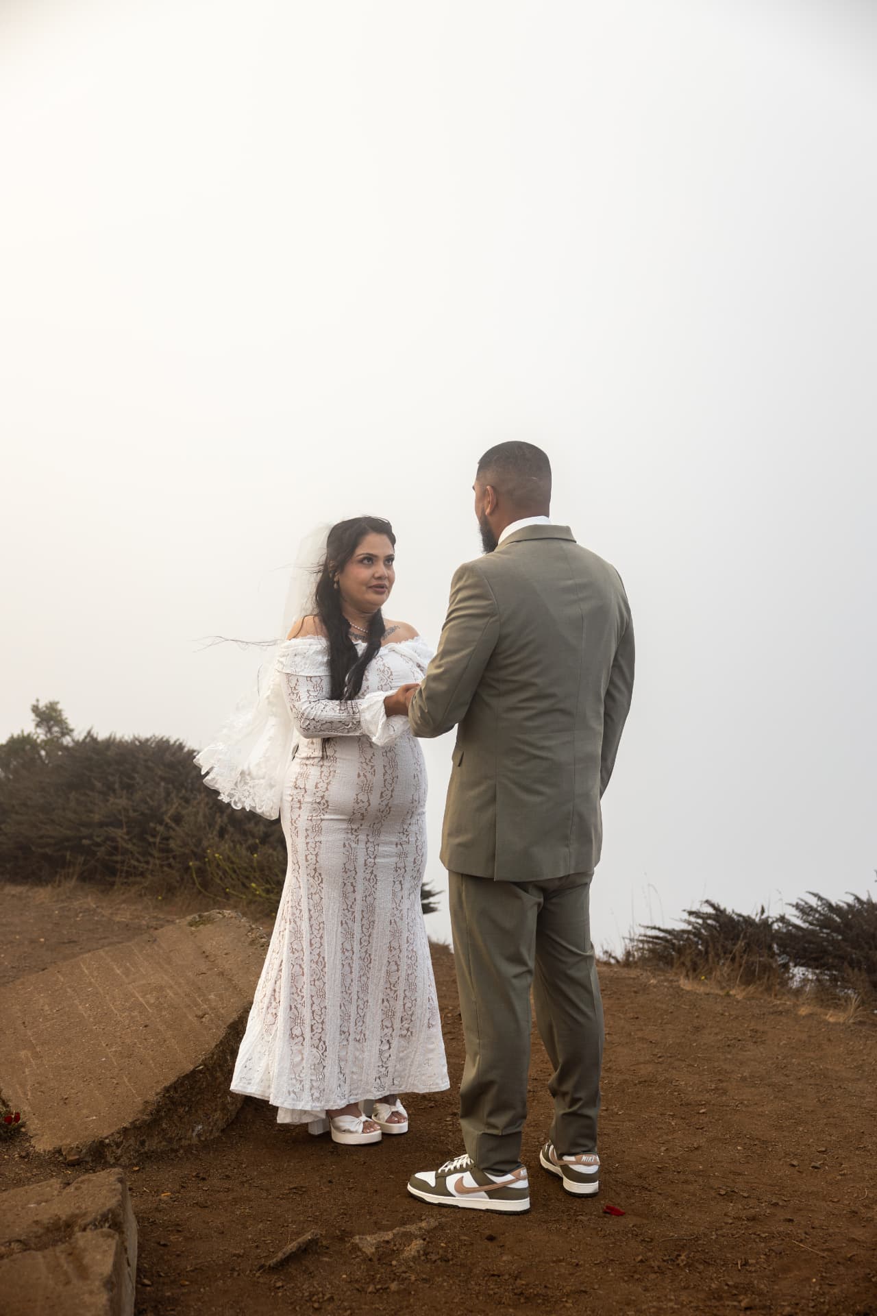 Bride in a white lace gown and groom in an olive-green suit holding hands facing each other on a foggy hilltop, Crystal & Joey wedding
