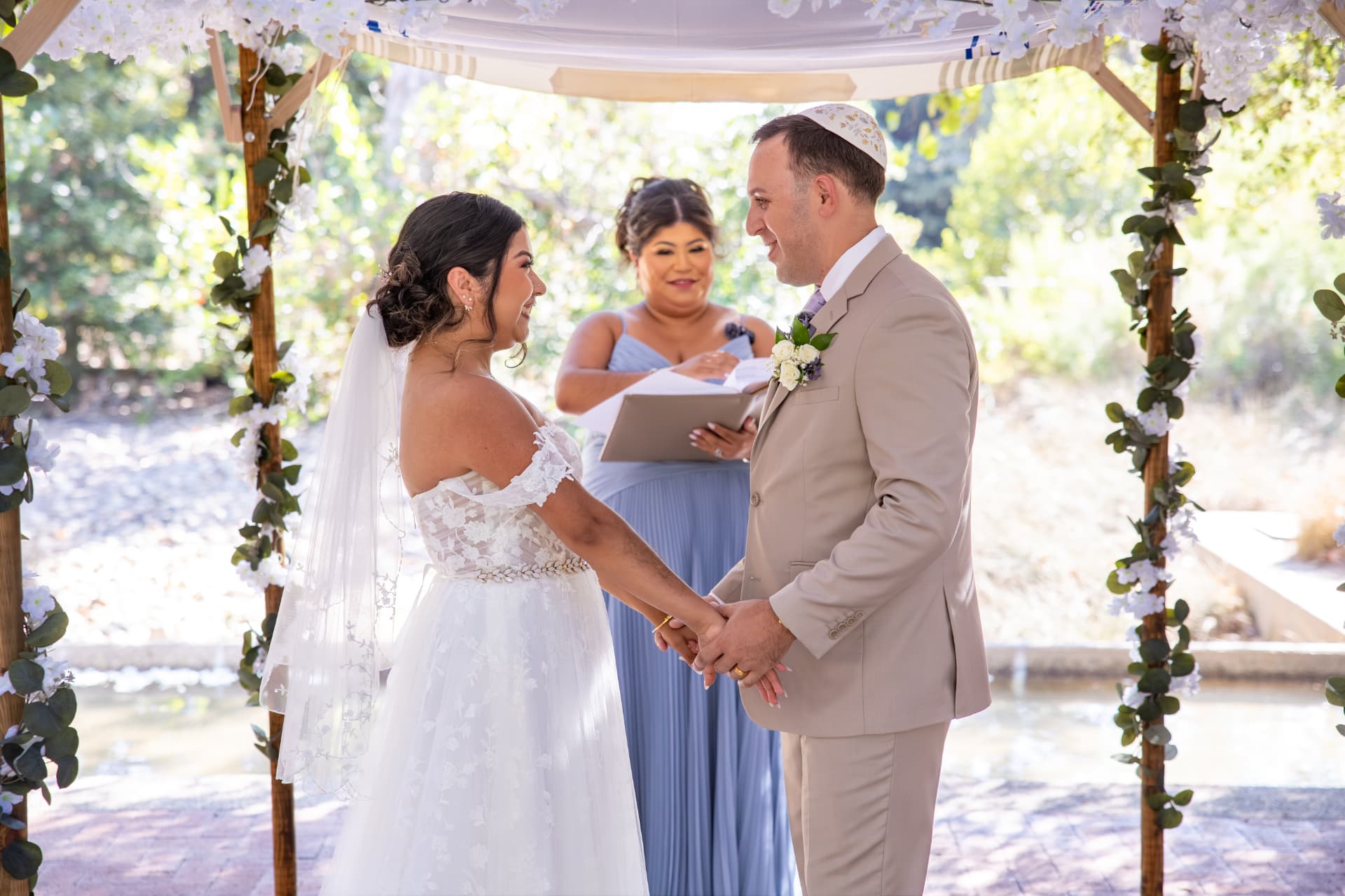 Ashley and Sean gazing at each other with joyful intimacy as they hold hands under a floral chuppah during their sunlit outdoor wedding ceremony
