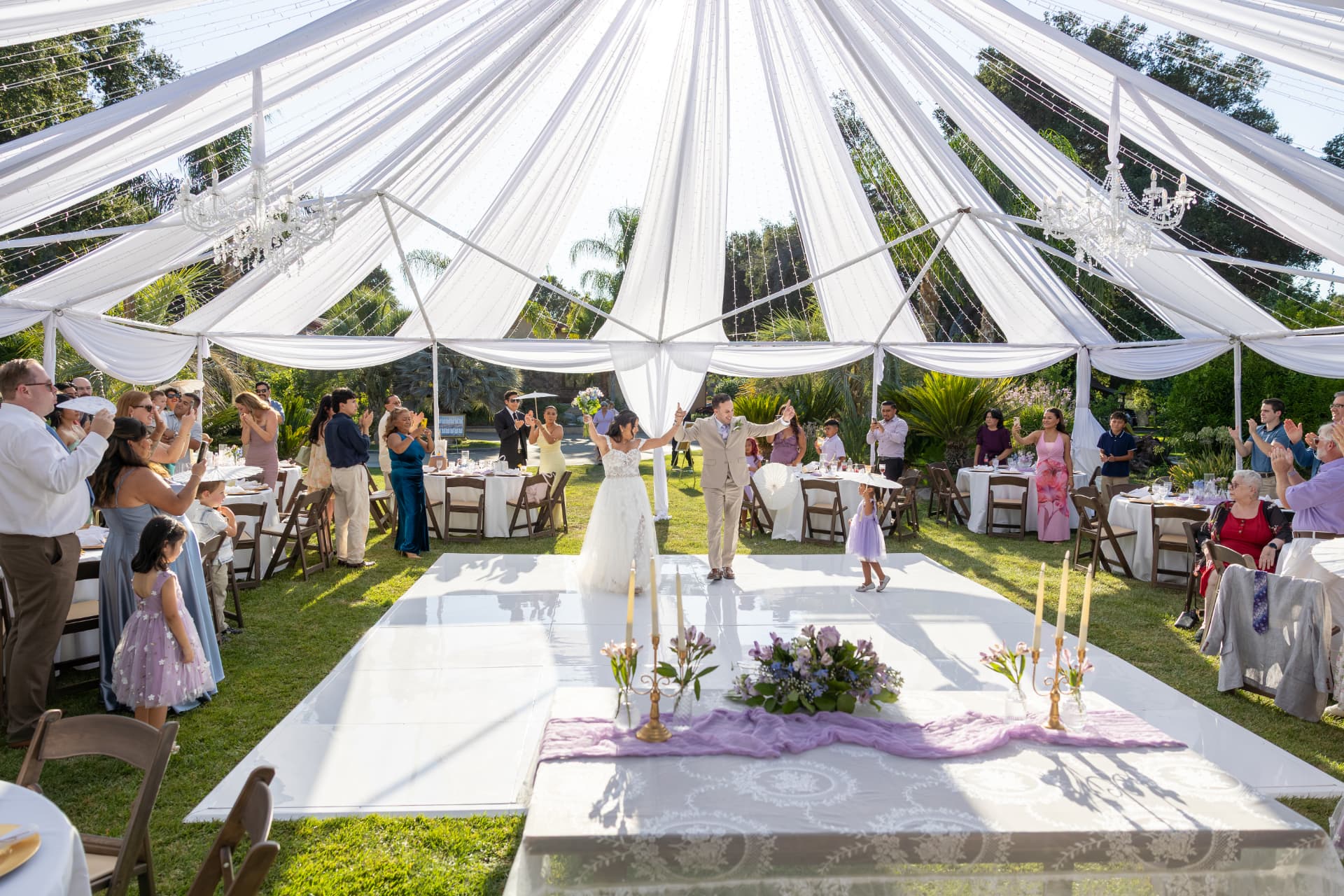 Bride and groom walking hand-in-hand onto a white dance floor under a draped white canopy with crystal chandeliers and string lights as guests cheer, Ashley & Sean wedding
