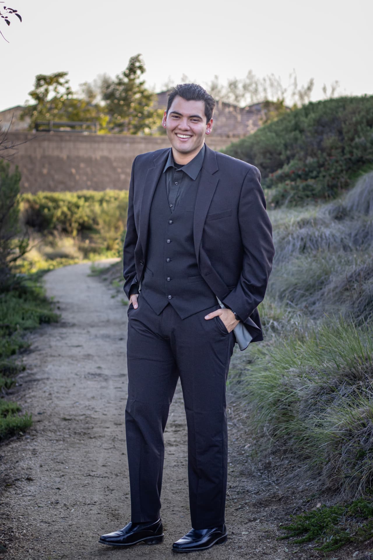 Abel Padilla in a charcoal three-piece suit standing confidently on an outdoor dirt path, smiling warmly in soft natural light