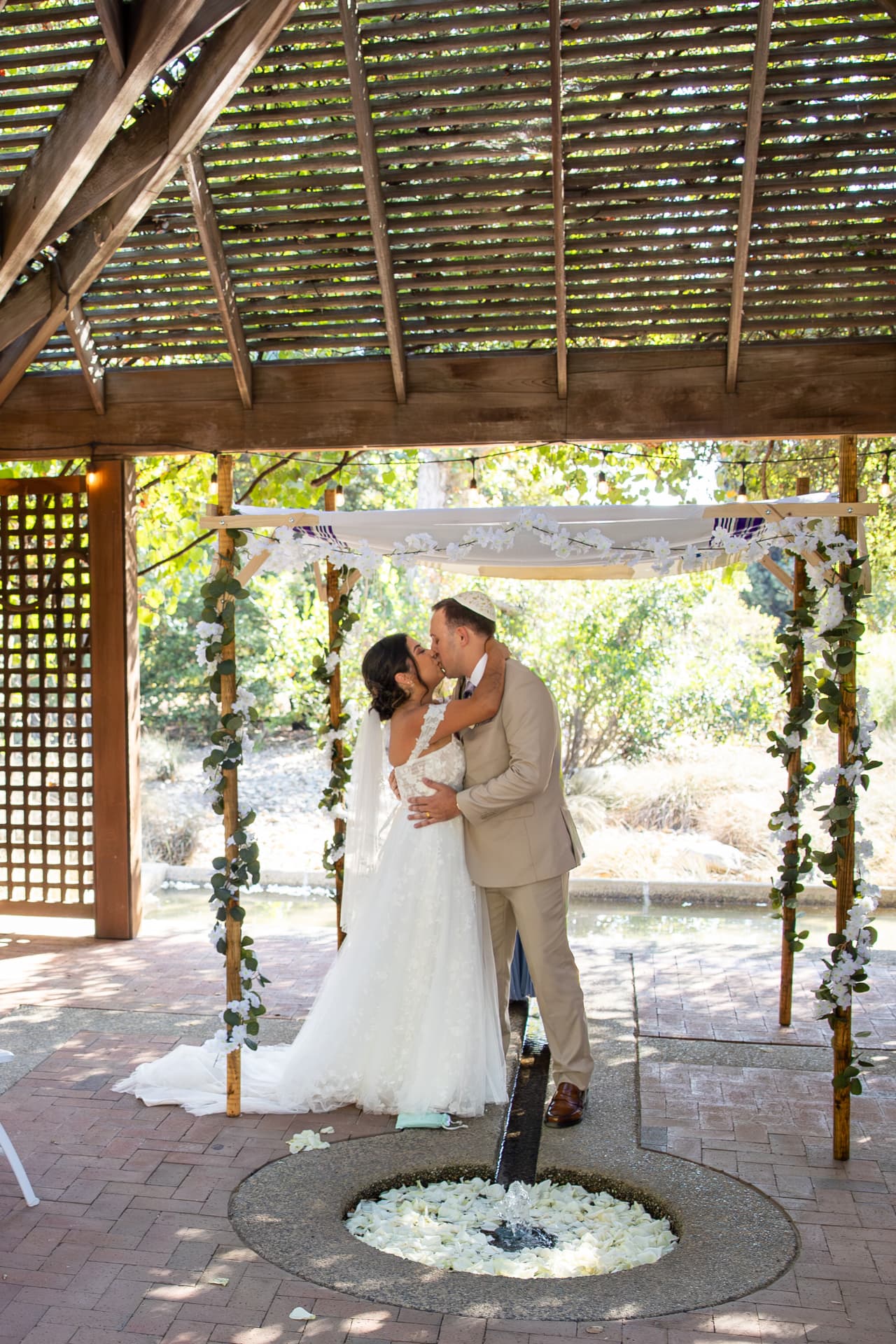 Ashley and Sean sharing a romantic first kiss under a white-flowered chuppah in a serene garden setting with a small stone fountain