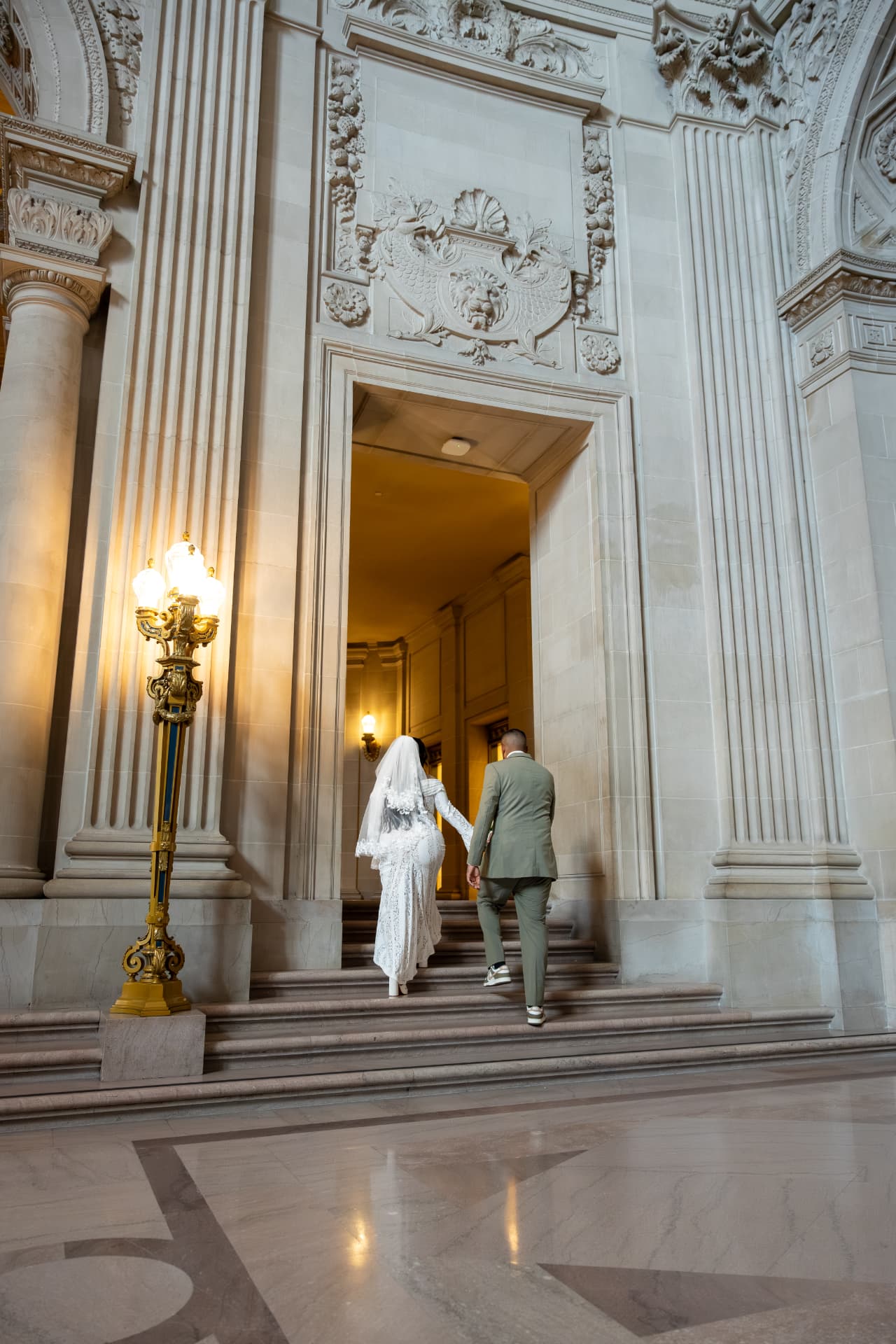 Bride in a white lace gown and groom in a sage-green suit walking up stone stairs toward a warmly lit doorway in a grand neoclassical hall, Crystal & Joey wedding