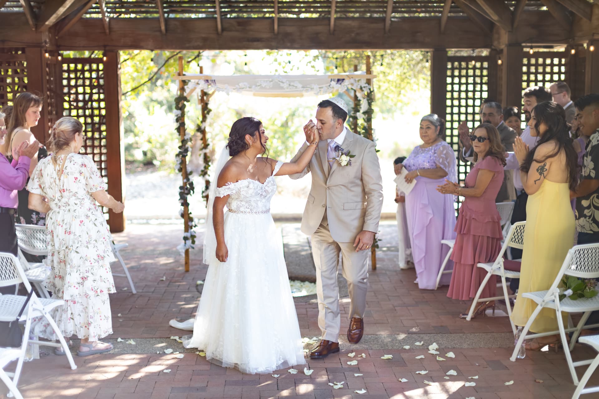 Ashley and Sean walking down the aisle in a celebratory mood as guests applaud their union in a sun-drenched, pergola-shaded garden