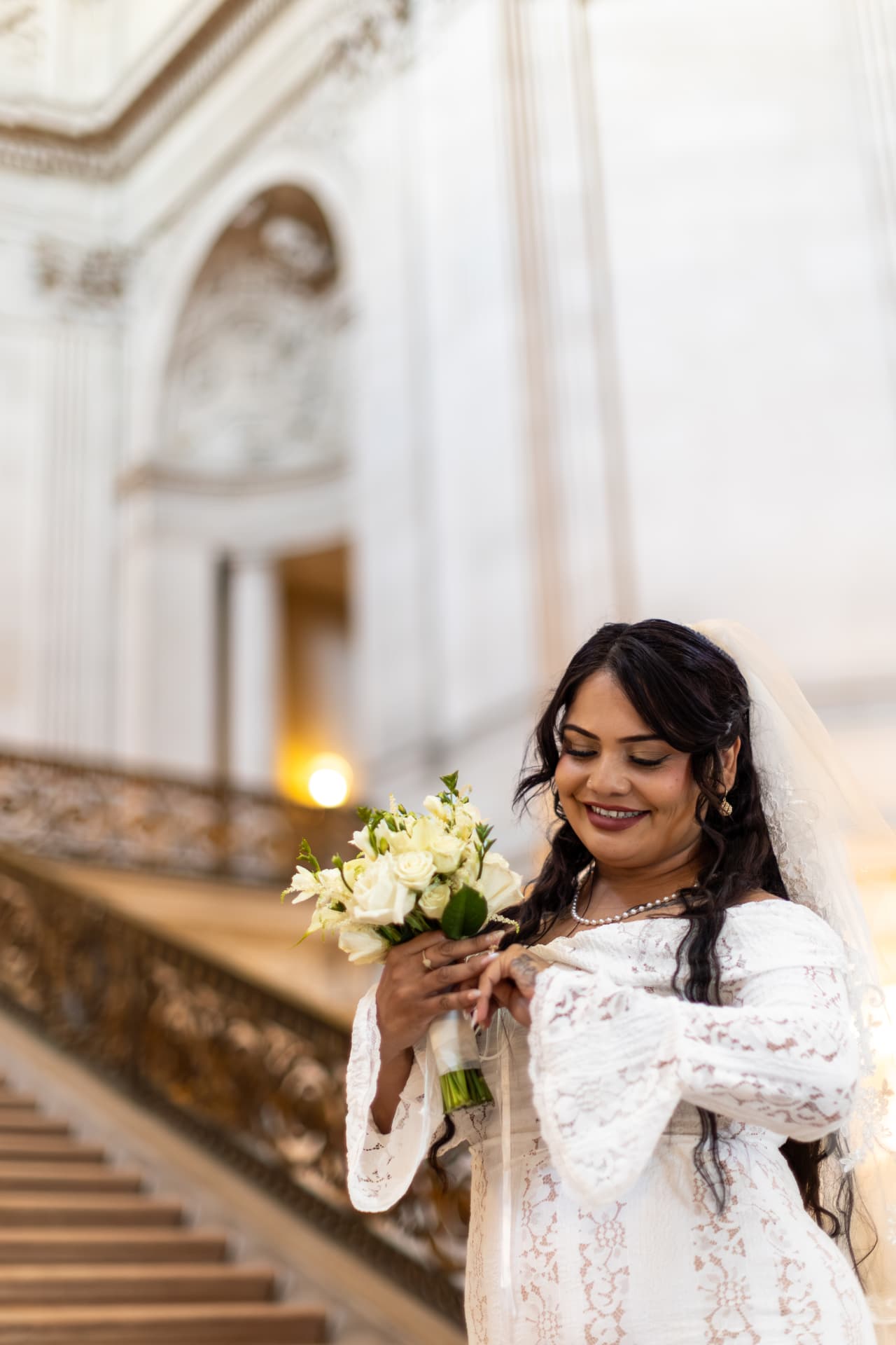 Bride looking down at her white rose bouquet with a soft smile in a brightly lit classical interior, wearing a lace dress with bell sleeves and a veil, Crystal & Joey wedding