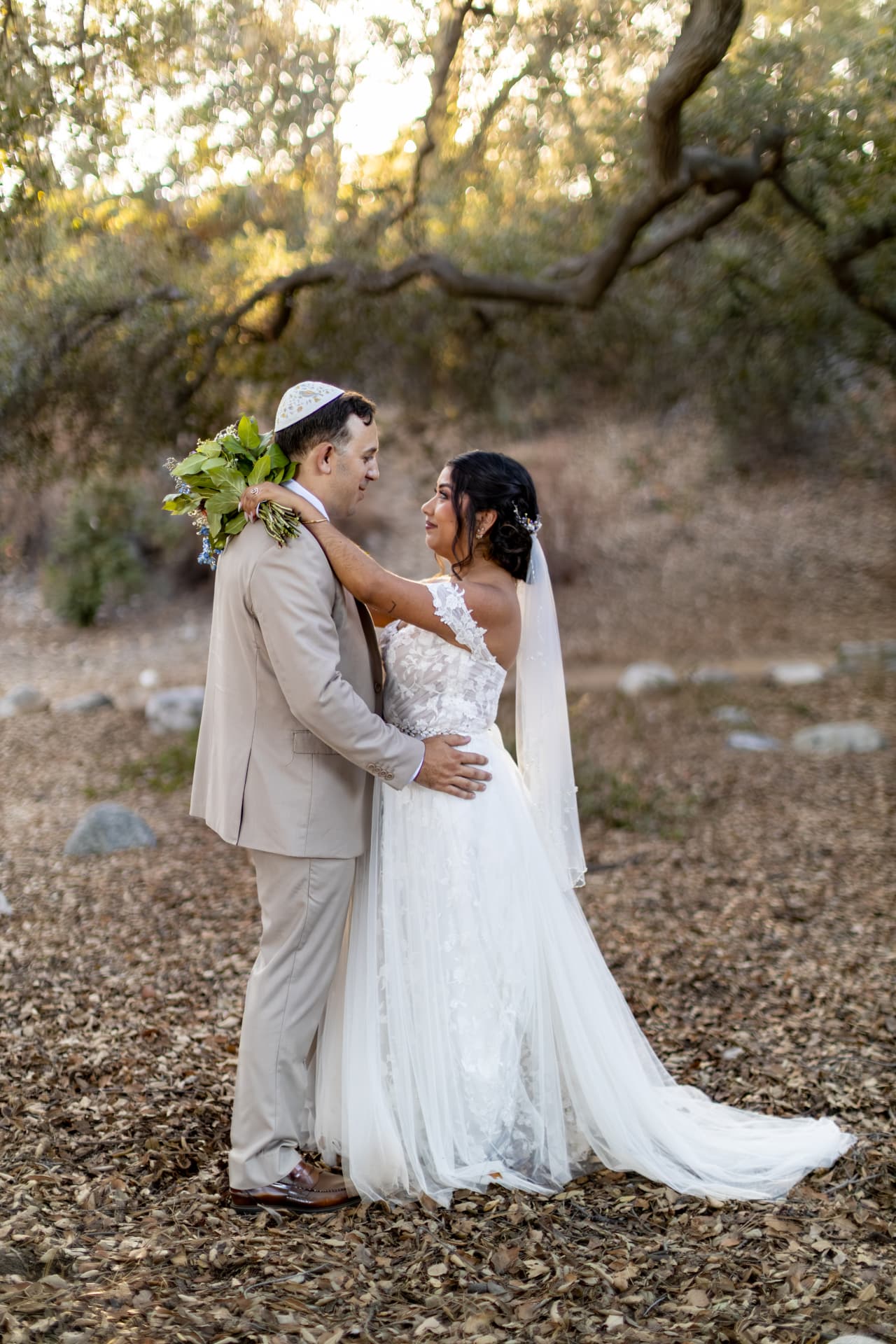 Bride and groom facing each other beneath sprawling oak branches, her train spread across the leaves