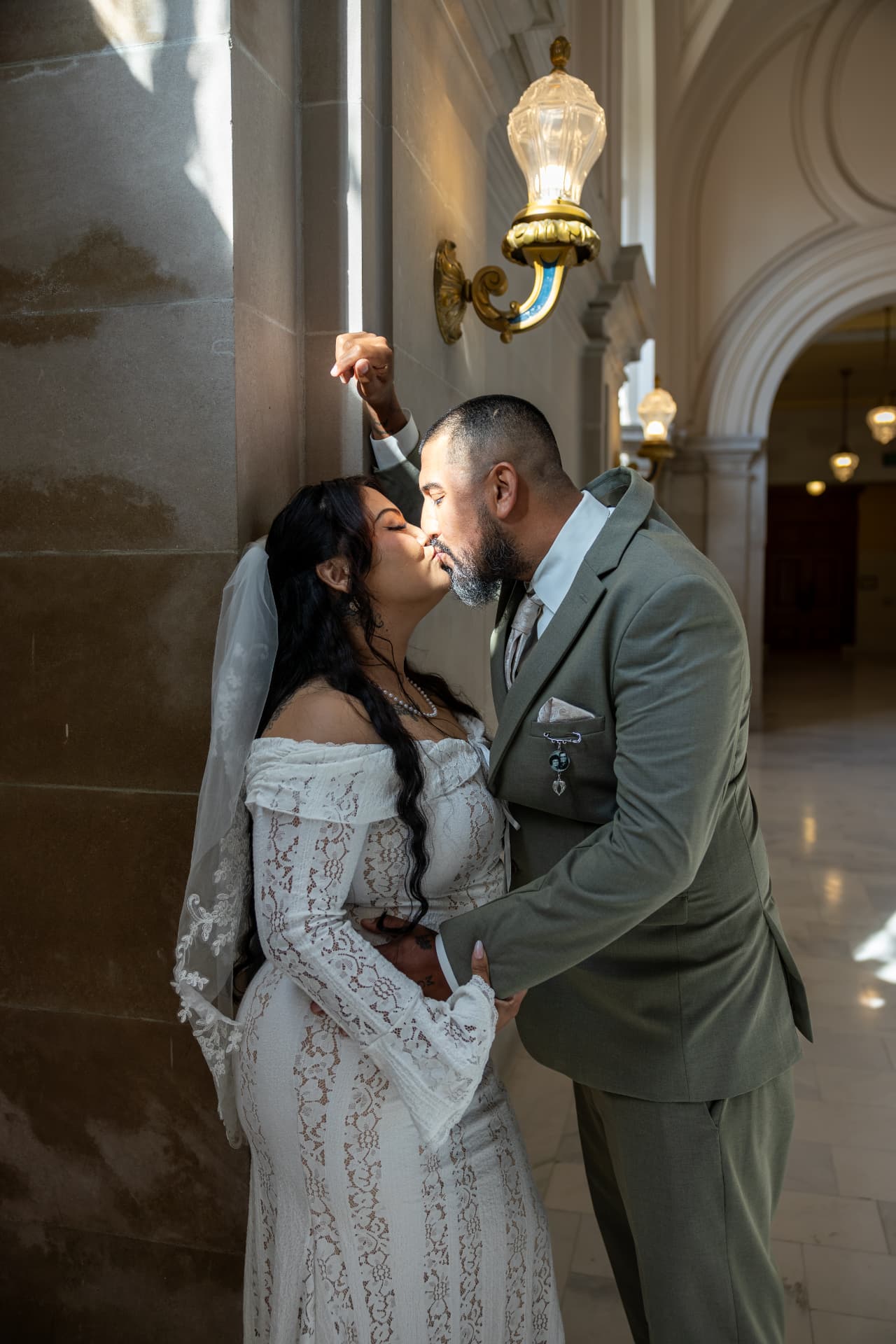 Bride and groom sharing an intimate kiss against a sunlit stone wall beside an ornate gold sconce inside a grand building, Crystal & Joey wedding