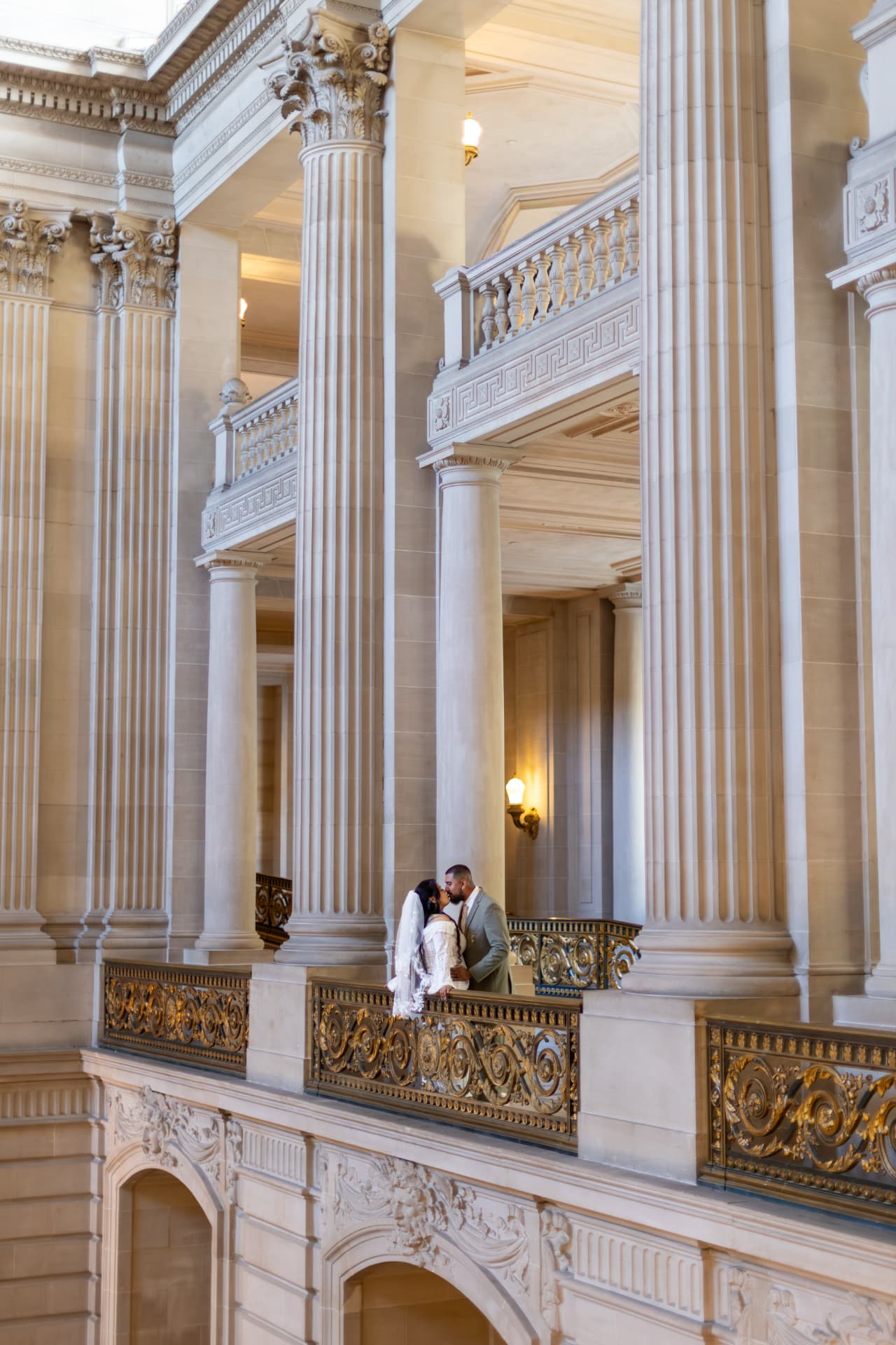 Bride and groom kissing on an upper balcony framed by massive fluted stone columns and an ornate gold railing, small within the grand classical architecture, Crystal & Joey wedding