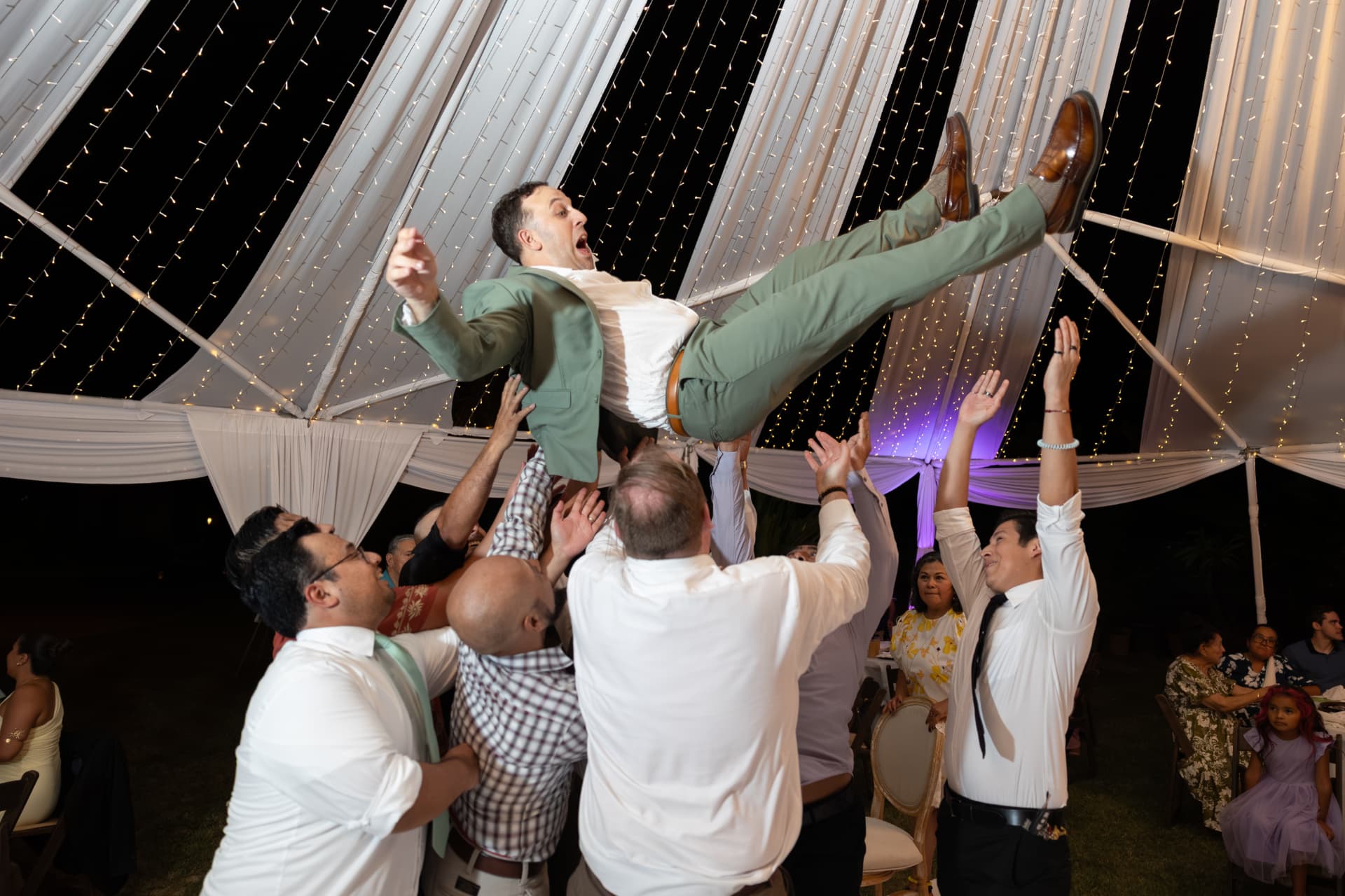 Sean tossed joyfully into the air by wedding guests under a canopy of white lights inside a large tent at the Ashley & Sean wedding reception