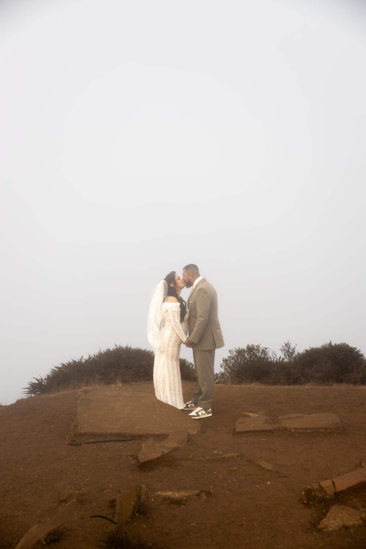 Bride in a white lace dress and groom in a tan suit kissing on a foggy dirt hilltop in soft diffused light, Crystal & Joey wedding
