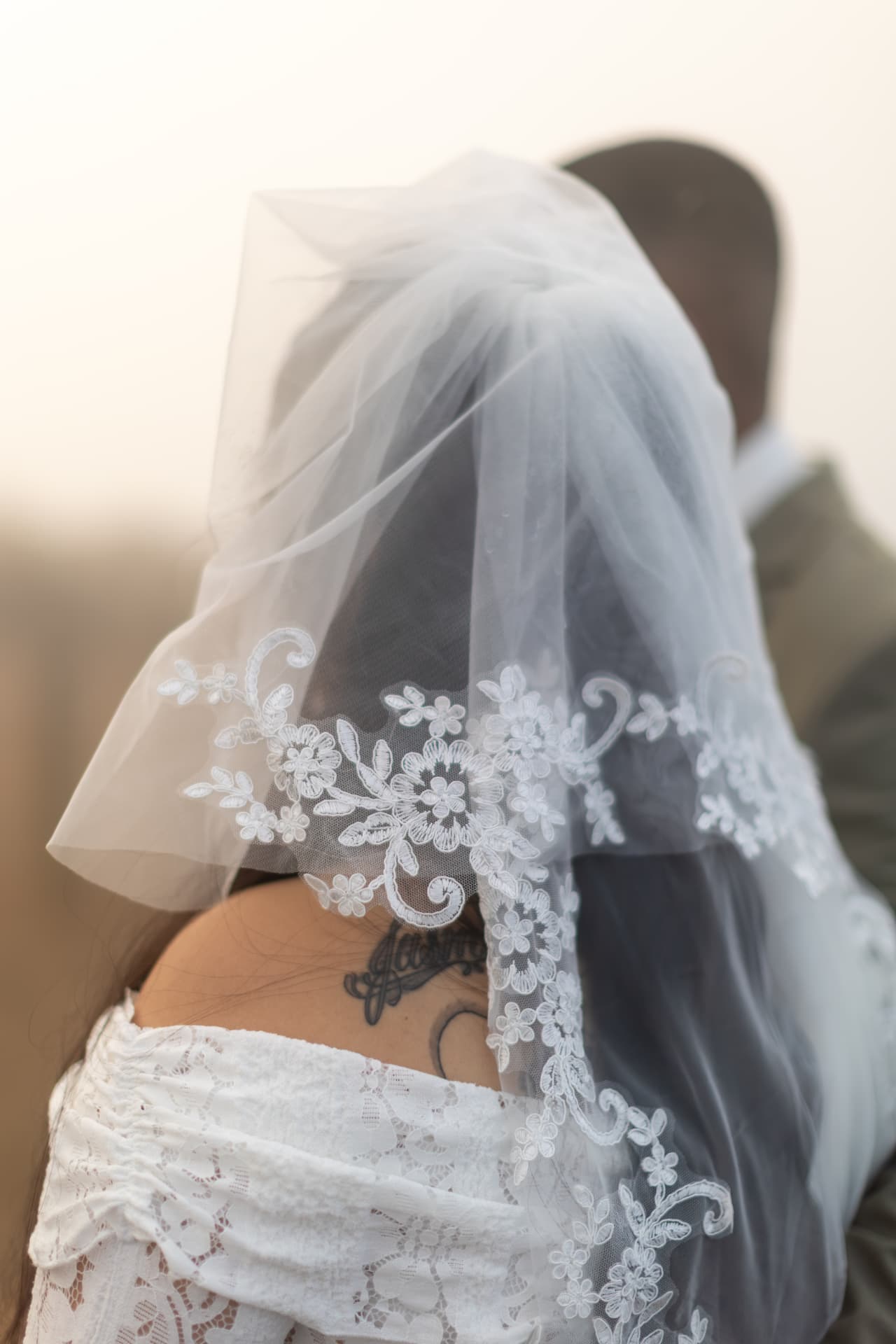 Bride seen from behind showing the floral lace embroidery of her veil and a crescent moon tattoo on her shoulder, the groom softly blurred in the hazy outdoor light, Crystal & Joey wedding