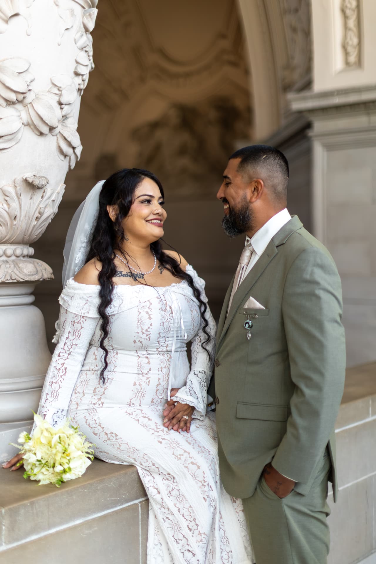 Bride in an off-the-shoulder lace gown seated on a stone ledge holding white and yellow flowers, smiling at the groom in an olive-green suit who holds her hand in a grand classical interior, Crystal & Joey wedding